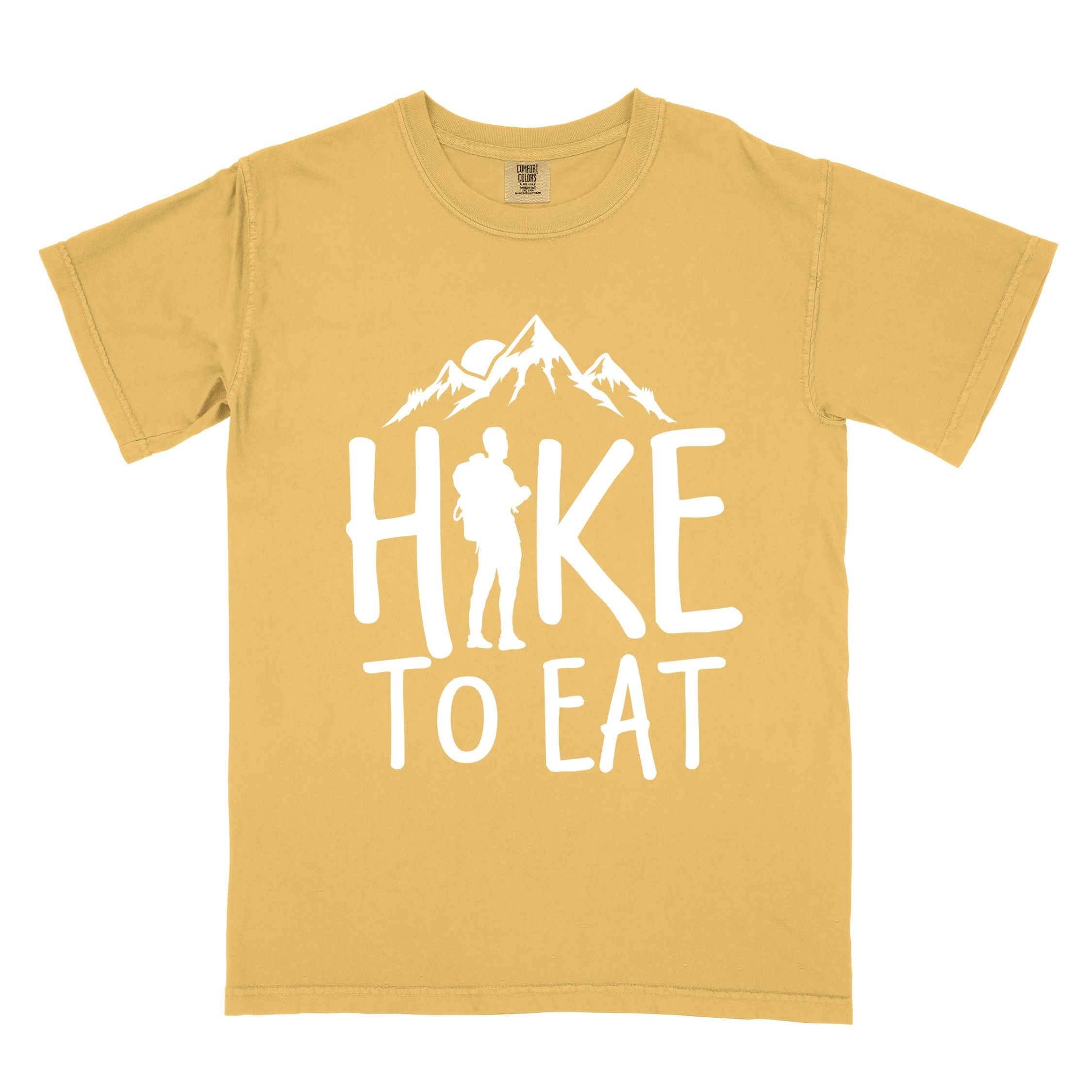 Mustard "Hike to Eat" colored statement tee with bold typography, mountains, birds, sunrise, perfect for food motivated individuals.