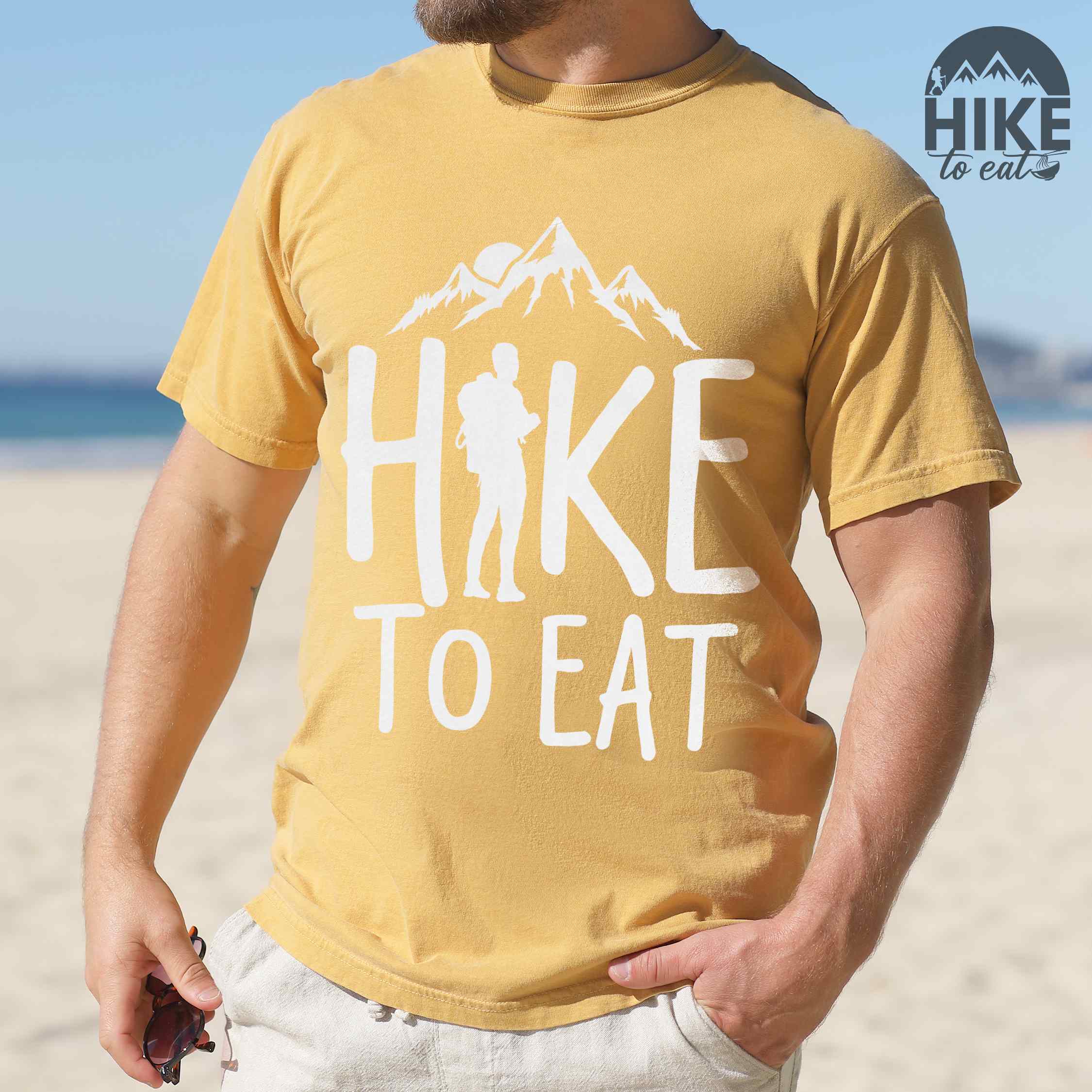 Man wearing mustard "Hike to Eat" colored statement tee with bold typography, mountains, birds, sunrise, perfect for food motivated individuals.
