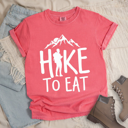 Pink shirt with a Hiking design featuring mountain silhouette and shows 'Hike To Eat' phrase in bold text