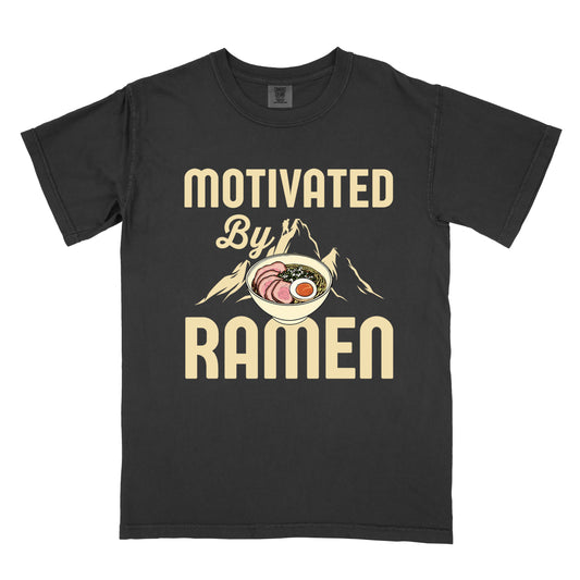 Black t-shirt with cream-colored text reading "MOTIVATED By RAMEN". "MOTIVATED" appears in large letters at the top, "By" in script in the middle, and "RAMEN" in large letters at the bottom. Features a detailed illustration of a ramen bowl with sliced meat, egg, and green onions in the center, positioned over cream-colored mountain silhouettes with a small hiker figure on a peak. 