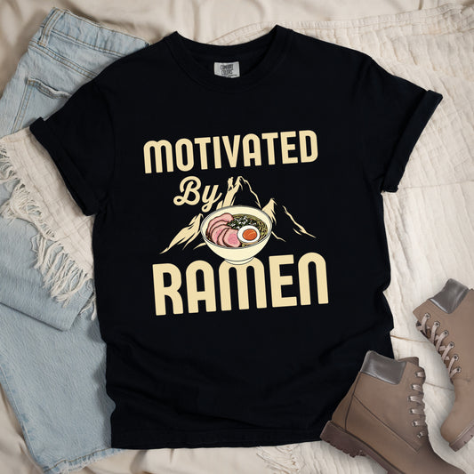 Black t-shirt with cream-colored text reading "MOTIVATED By RAMEN". "MOTIVATED" appears in large letters at the top, "By" in script in the middle, and "RAMEN" in large letters at the bottom. Features a detailed illustration of a ramen bowl with sliced meat, egg, and green onions in the center, positioned over cream-colored mountain silhouettes with a small hiker figure on a peak.  Features a classic crew neck and casual rolled sleeves, styled alongside light wash denim and brown hiking boots. 