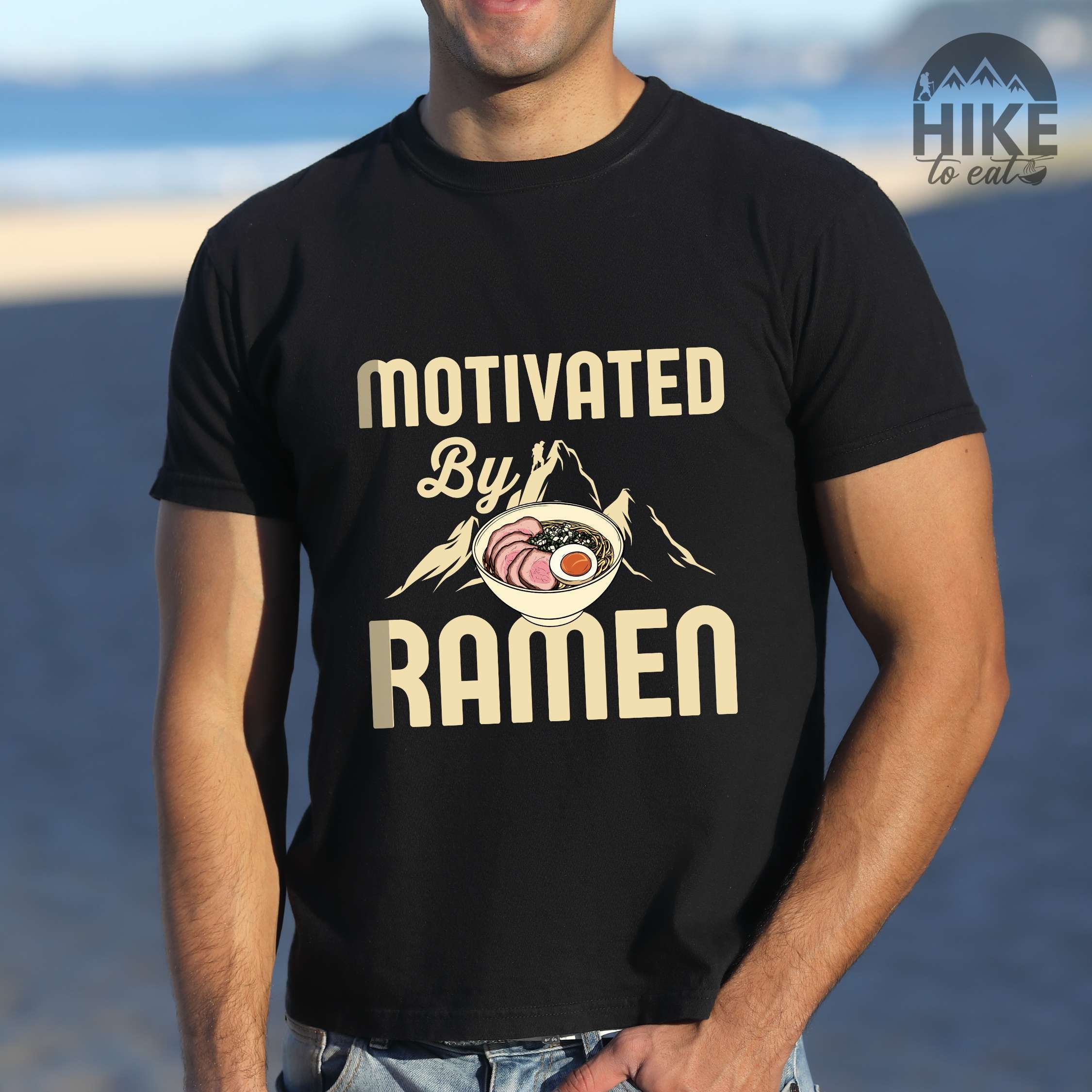 Man wearing black “Motivated by Ramen” colored vintage tee with bold text, mountain, Illustration of hot ramen. Perfect tee for foodie craving for a bowl of ramen,