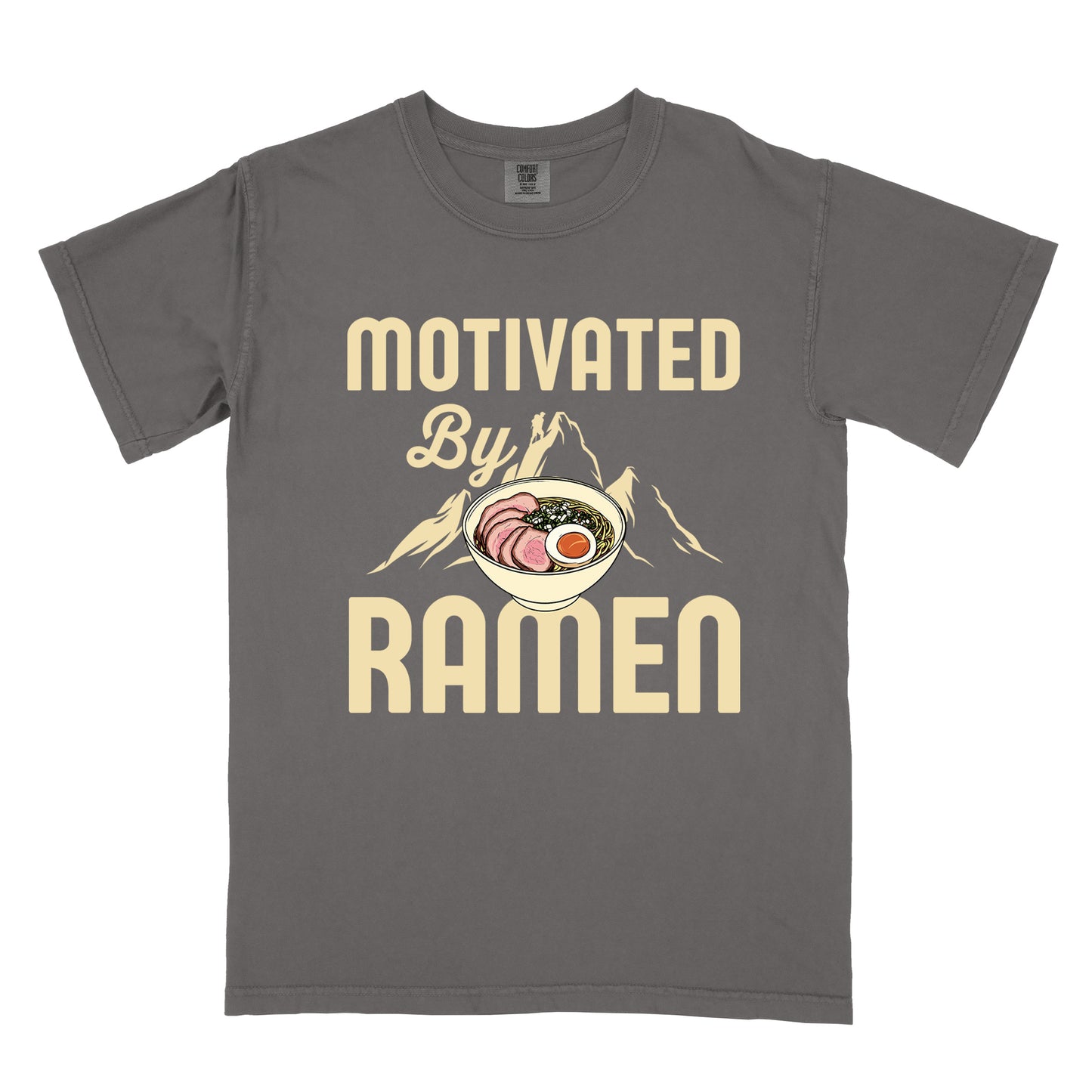 Pepper (Dark Gray) t-shirt with cream-colored text reading "MOTIVATED By RAMEN". "MOTIVATED" appears in large letters at the top, "By" in script in the middle, and "RAMEN" in large letters at the bottom. Features a detailed illustration of a ramen bowl with sliced meat, egg, and green onions in the center, positioned over cream-colored mountain silhouettes with a small hiker figure on a peak. 