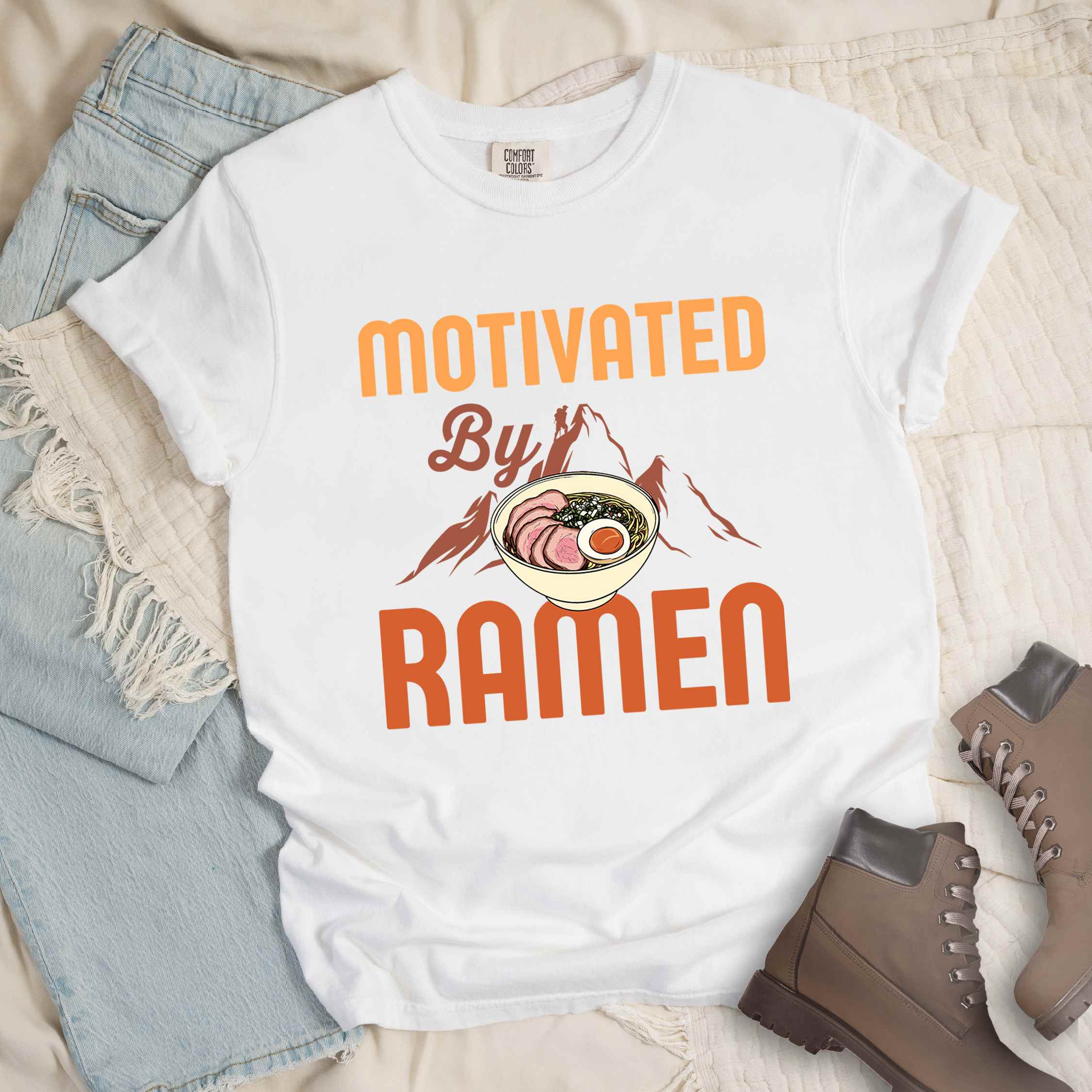 White “Motivated by Ramen” colored vintage tee with bold text, mountain, Illustration of hot ramen. Perfect tee for foodie craving for a bowl of ramen,