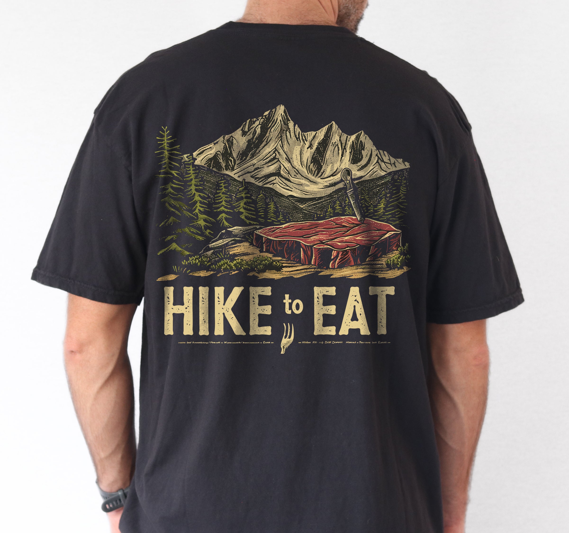 Black Hike to Eat t-shirt back view showing large design featuring raw steak in mountain wilderness landscape with pine trees worn by a male person.