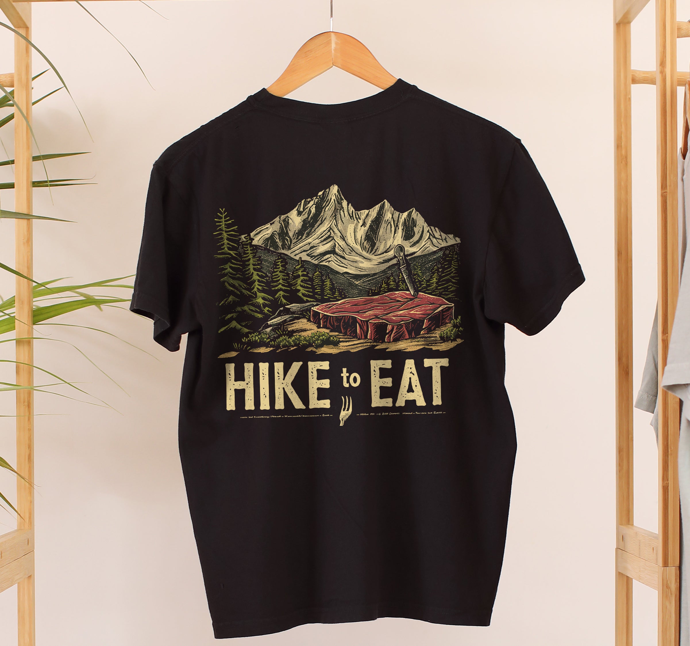 Hanging black Hike to Eat t-shirt back view showing large design featuring raw steak in mountain wilderness landscape with pine trees