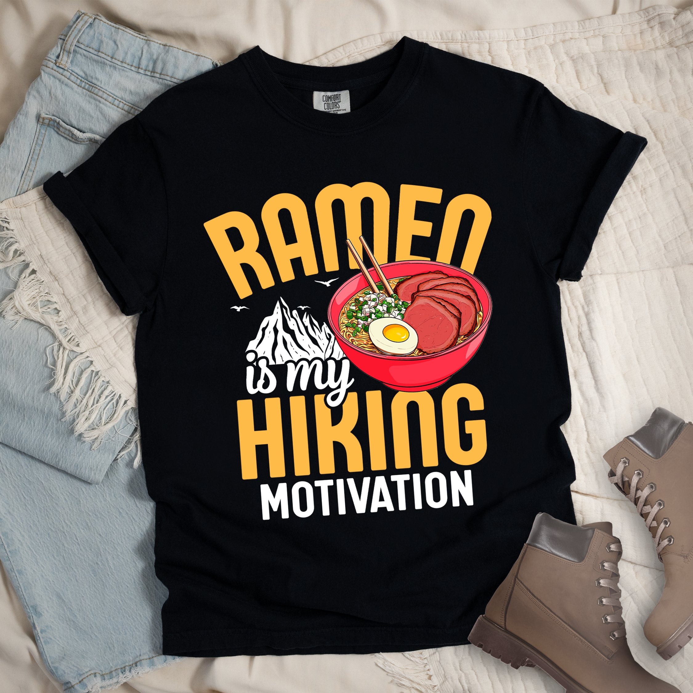 Black shirt with “Ramen is my Hiking Motivation” text, featuring a mountain graphic and a ramen bowl with noodles, egg, meat, and chopsticks.