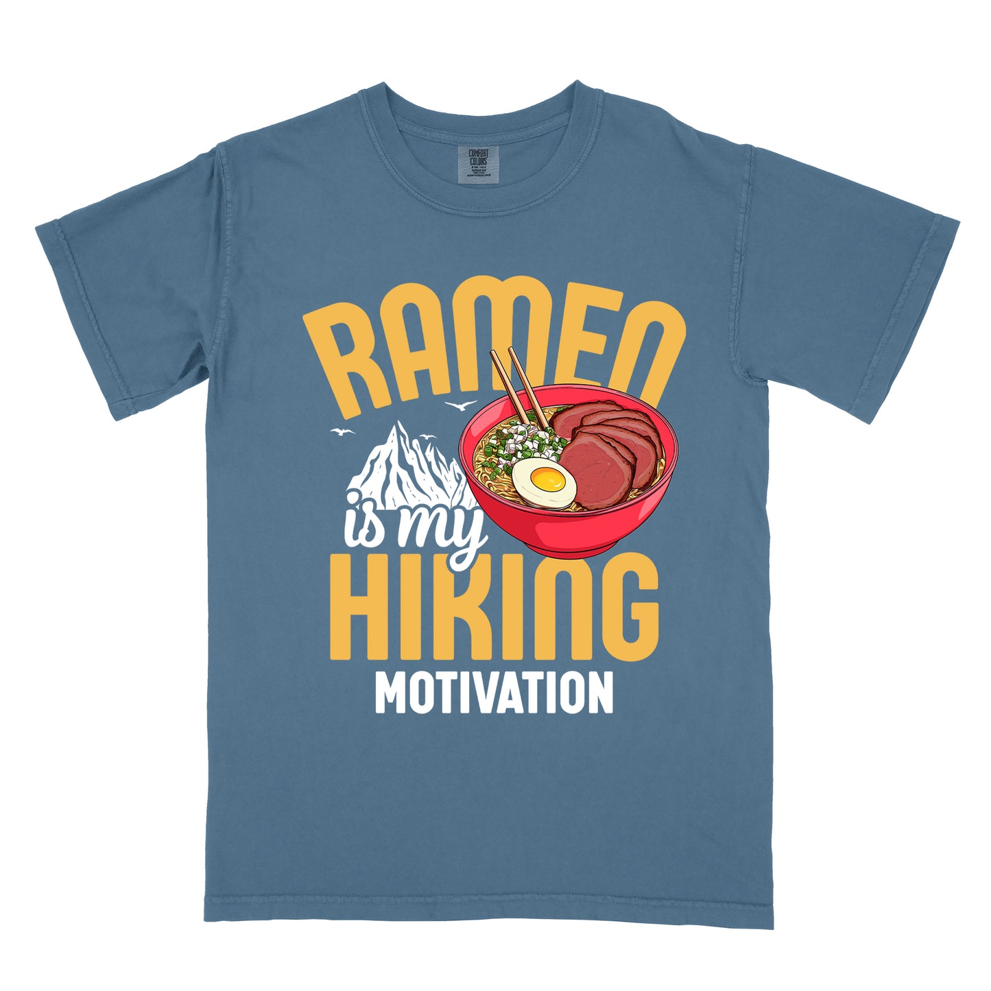 Blue Jean (Denim Blue) t-shirt with "RAMEN is my HIKING MOTIVATION" text design. "RAMEN" and "HIKING" appear in large yellow letters, with "is my" in white script and "MOTIVATION" in white capitals. The design features a colorful illustration of a red ramen bowl with sliced meat, egg, green onions, and chopsticks. A white mountain silhouette appears between "RAMEN" and "HIKING", with small bird silhouettes nearby.