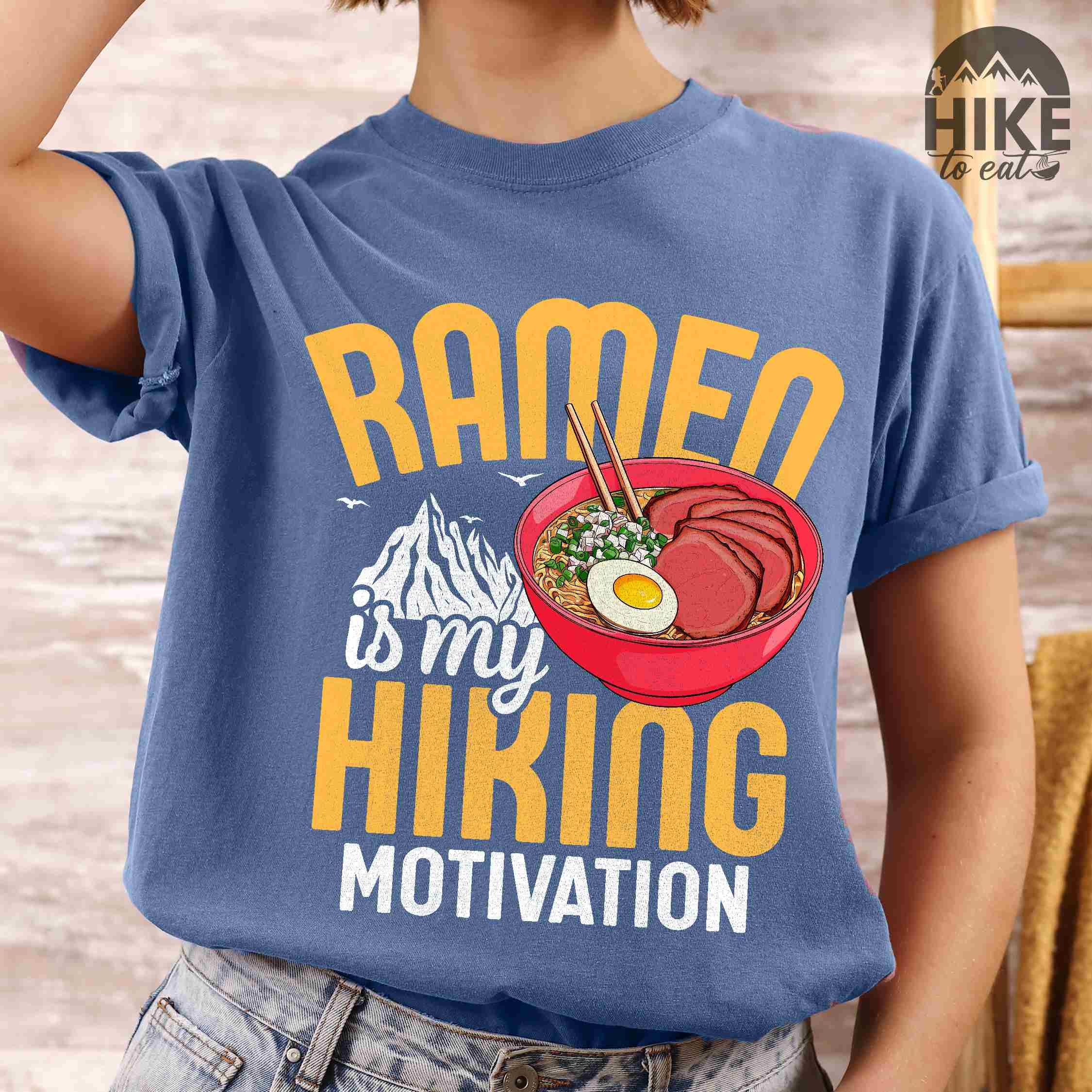 Woman wearing blue jean “Ramen is my Hiking motivation” colored vintage tee with bold text, mountain, Illustration of hot ramen. Perfect tee for foodie craving for a bowl of ramen,