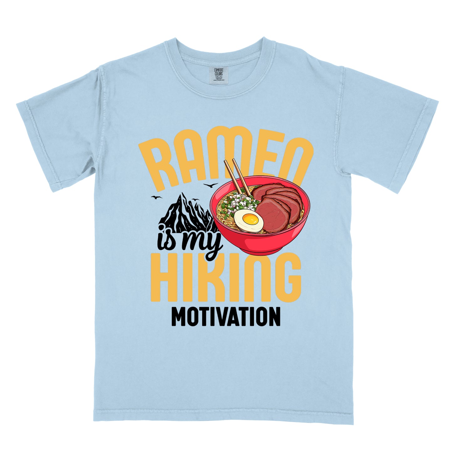 Chambray (Light Blue) t-shirt with "RAMEN is my HIKING MOTIVATION" text design. "RAMEN" and "HIKING" appear in large yellow letters, with "is my" in black script and "MOTIVATION" in black capitals. The design features a colorful illustration of a red ramen bowl with sliced meat, egg, green onions, and chopsticks. A black mountain silhouette appears between "RAMEN" and "HIKING", with small bird silhouettes nearby.