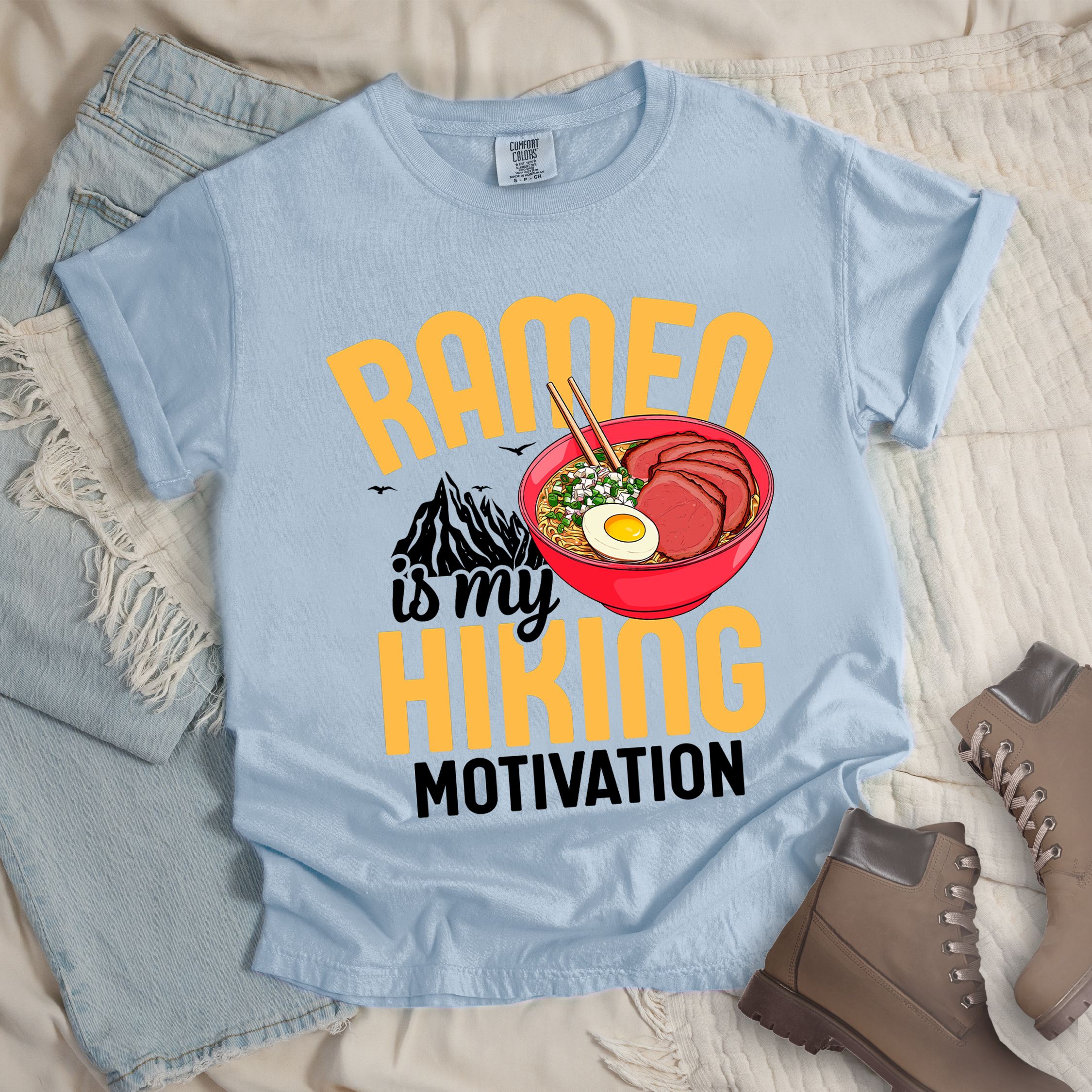 Light blue shirt with “Ramen is my Hiking Motivation” text, featuring a mountain graphic and a ramen bowl with noodles, egg, meat, and chopsticks.