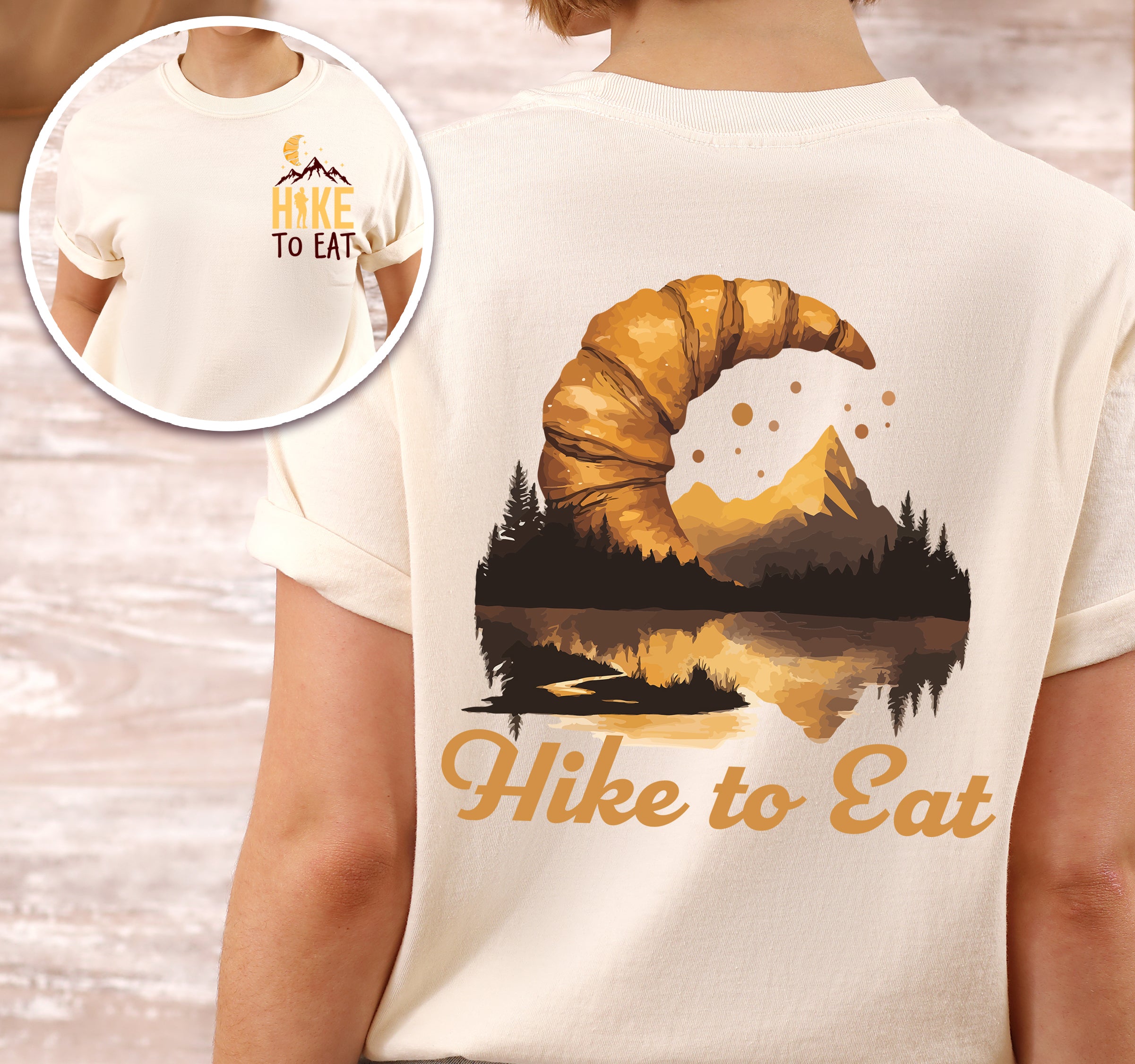 Ivory (Cream White) Hike to Eat t-shirt front and back view showing small chest logo and large back design featuring golden croissant filled with sunset mountain lake landscape, pine trees, and reflected peaks.