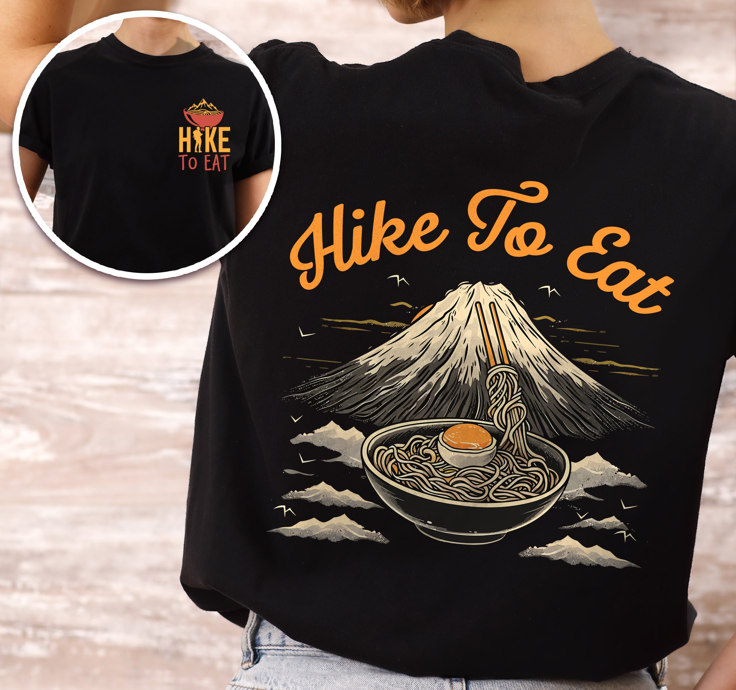 Black Hike to Eat t-shirt front and back view showing small orange chest logo with ramen bowl and large back design featuring black steaming ramen bowl with swirling noodles and soft-boiled egg in front of sketched Mount Fuji landscape with clouds.
