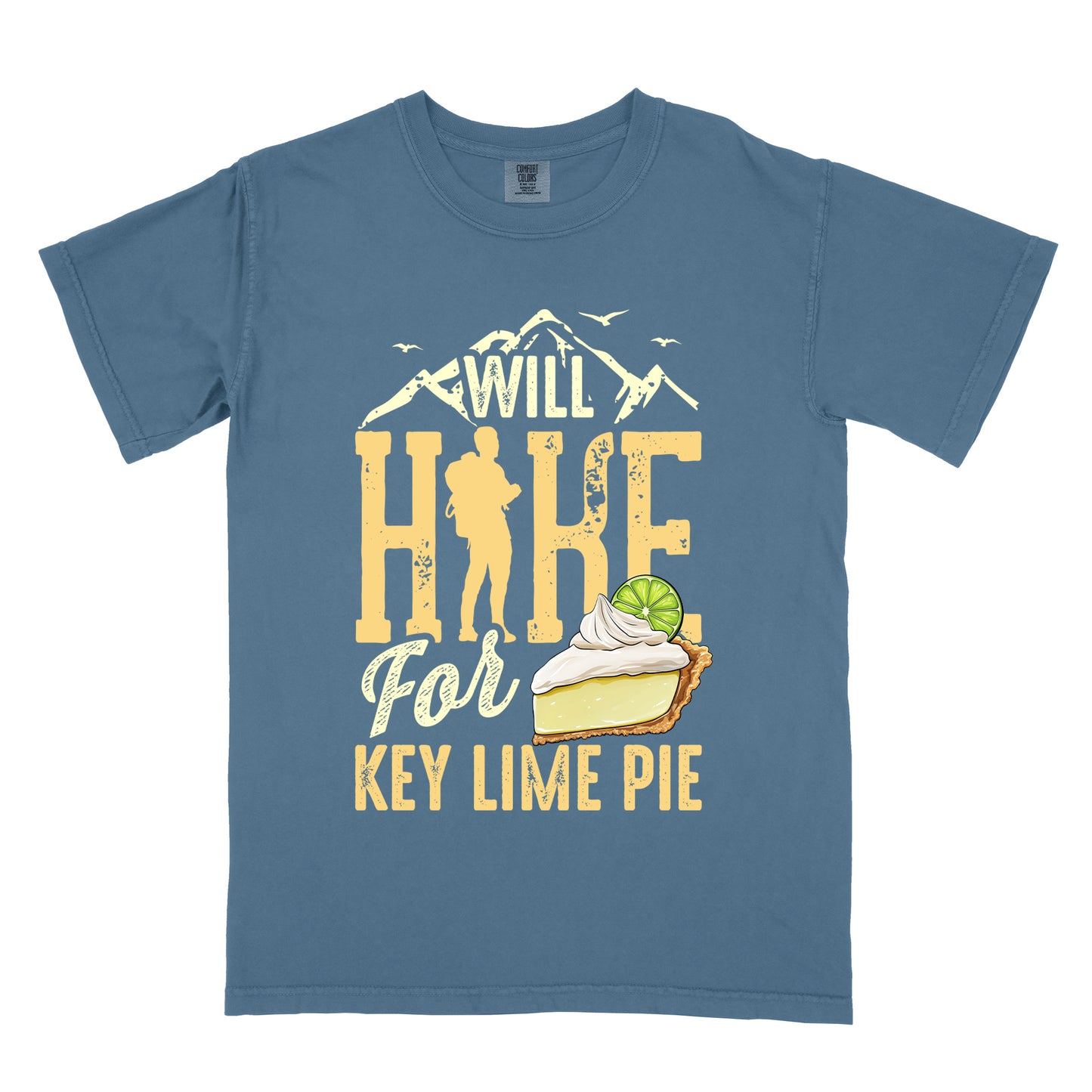Blue Jean (Denim Blue) t-shirt with text "WILL HIKE For KEY LIME PIE" in yellow tones. "WILL" appears in cream letters within mountain silhouettes with flying birds. "HIKE" is in large distressed yellow letters with a hiker silhouette replacing the "I". "For" appears in cream script, and "KEY LIME PIE" in yellow block letters. An illustration of a key lime pie slice with whipped cream and lime garnish appears next to "For" and above "KEY LIME PIE".