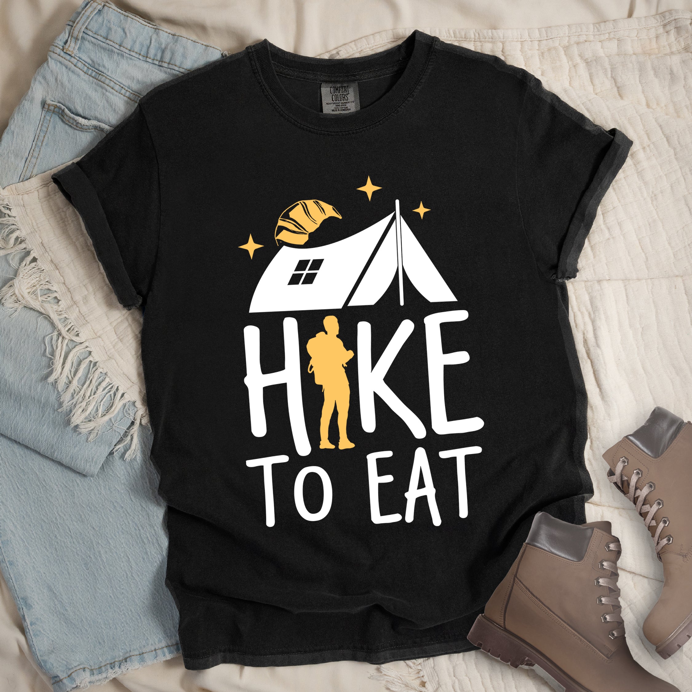 Black graphic t-shirt with 'Hike to Eat' text featuring white camping tent, yellow hiker silhouette, croissant, and stars design, styled with light wash jeans and brown hiking boots