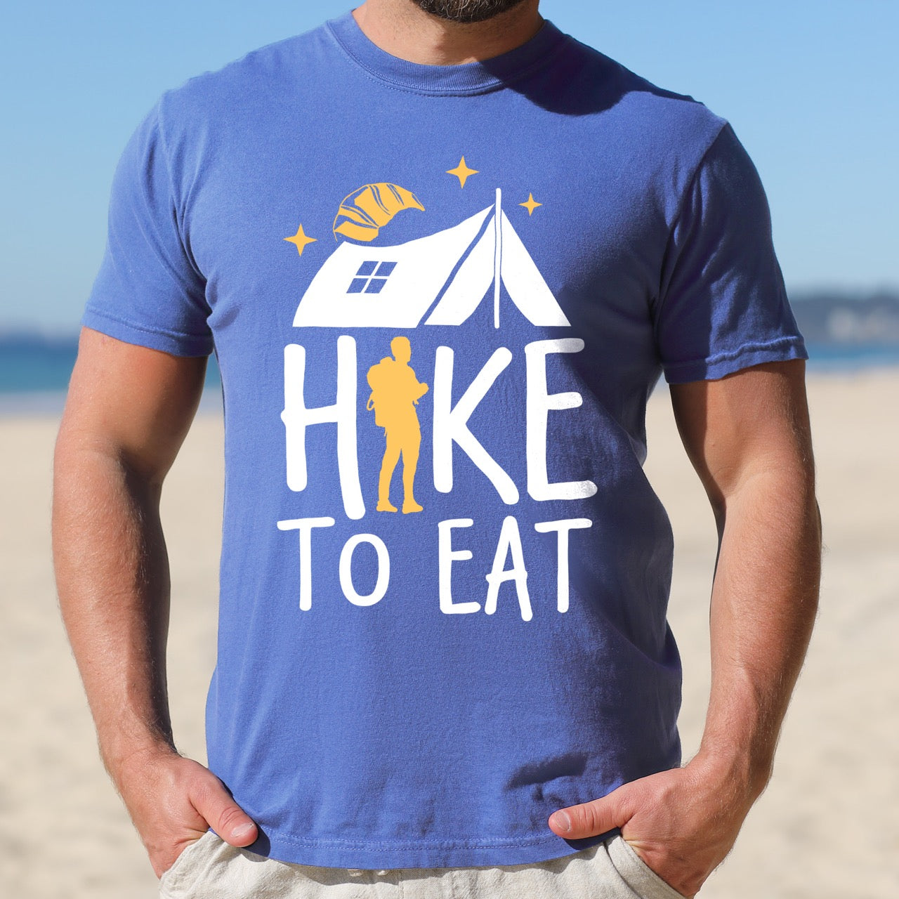 Man wearing flo blue Hike to Eat brand philosophy vintage tee with white camping tent croissant moon stars character silhouette with backpack integrated into typography on Comfort Colors shirt for food motivated adventurers, outdoor enthusiasts and hikers