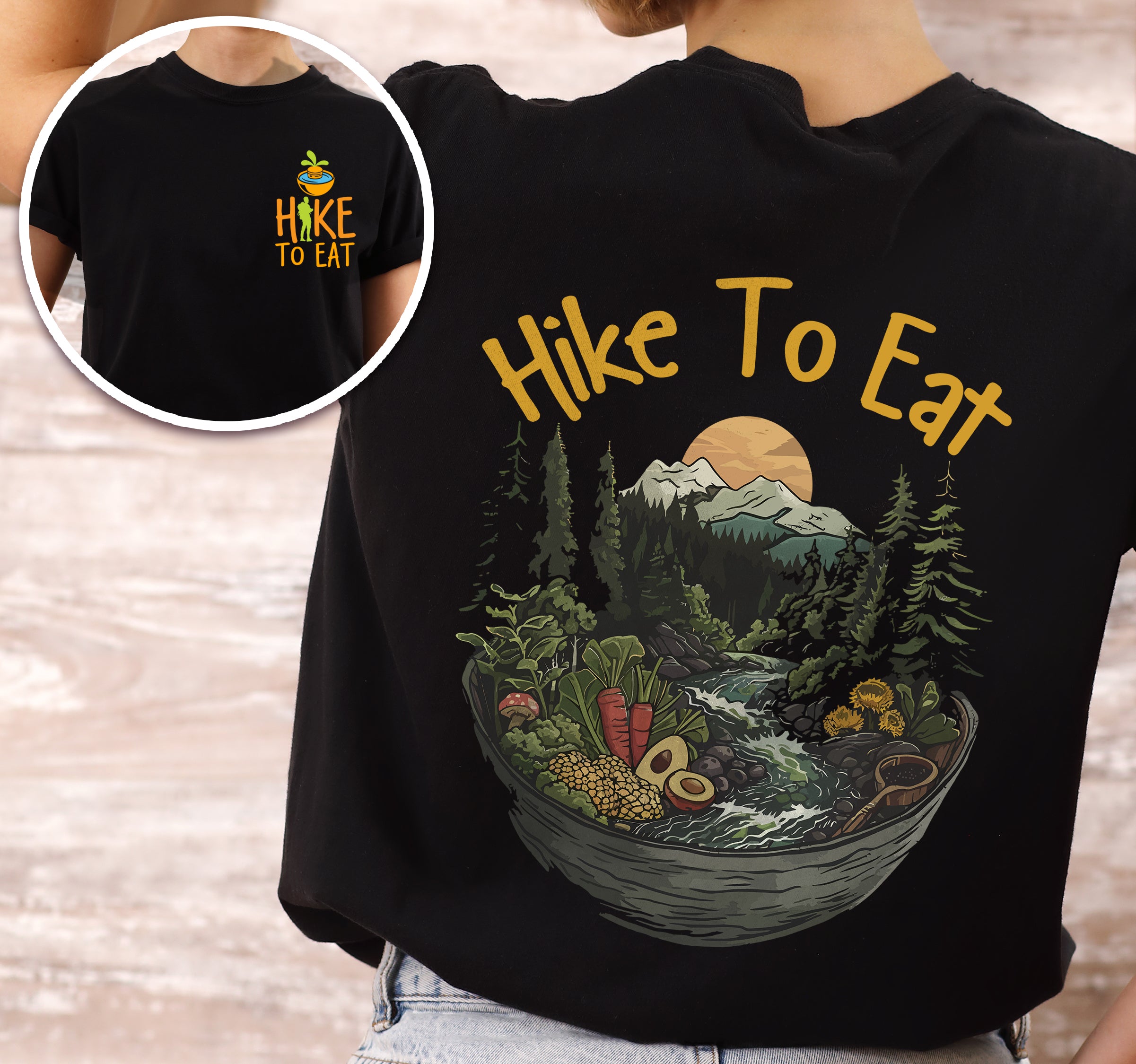 Black Hike to Eat t-shirt front and back view showing small chest design and large back design featuring bowl-shaped mountain landscape with pine trees, waterfall, sunset, and various fresh foods including avocado and vegetables worn by a woman.