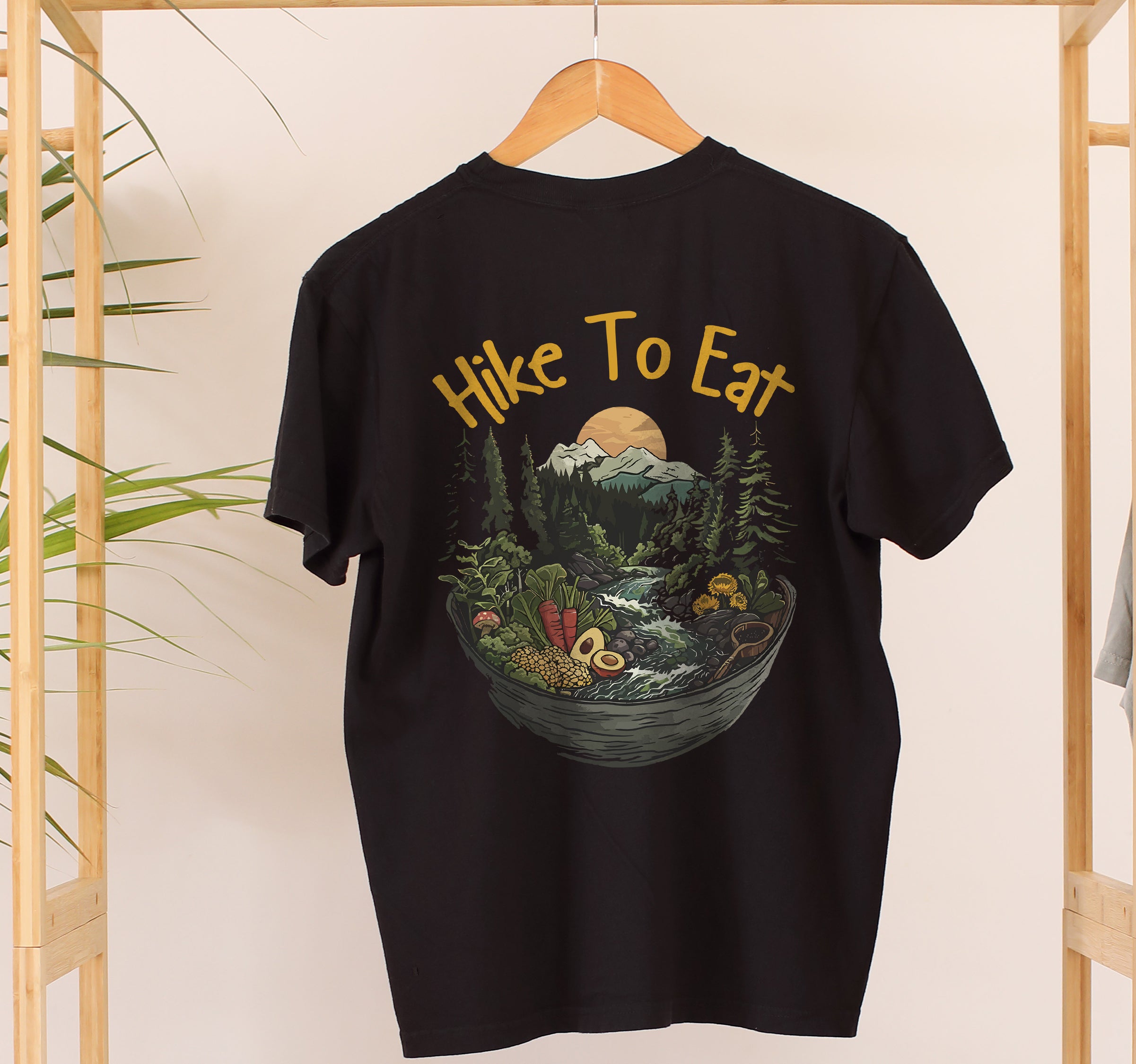 Black Hike to Eat t-shirt on a wooden hanger with back view showing large design featuring bowl-shaped mountain landscape with pine trees, waterfall, sunset, and various fresh foods including avocado and vegetables.