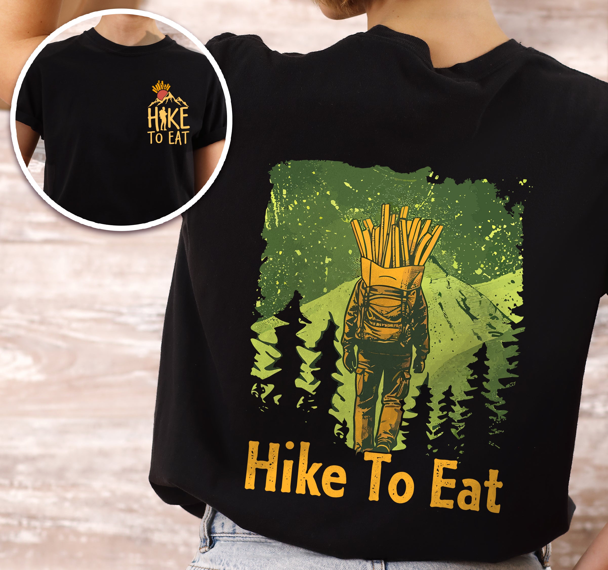 Black Hike to Eat t-shirt front and back view showing small french fries chest detail and large back design featuring hiker with french fries backpack walking through green forest landscape with pine tree silhouettes worn by a woman.