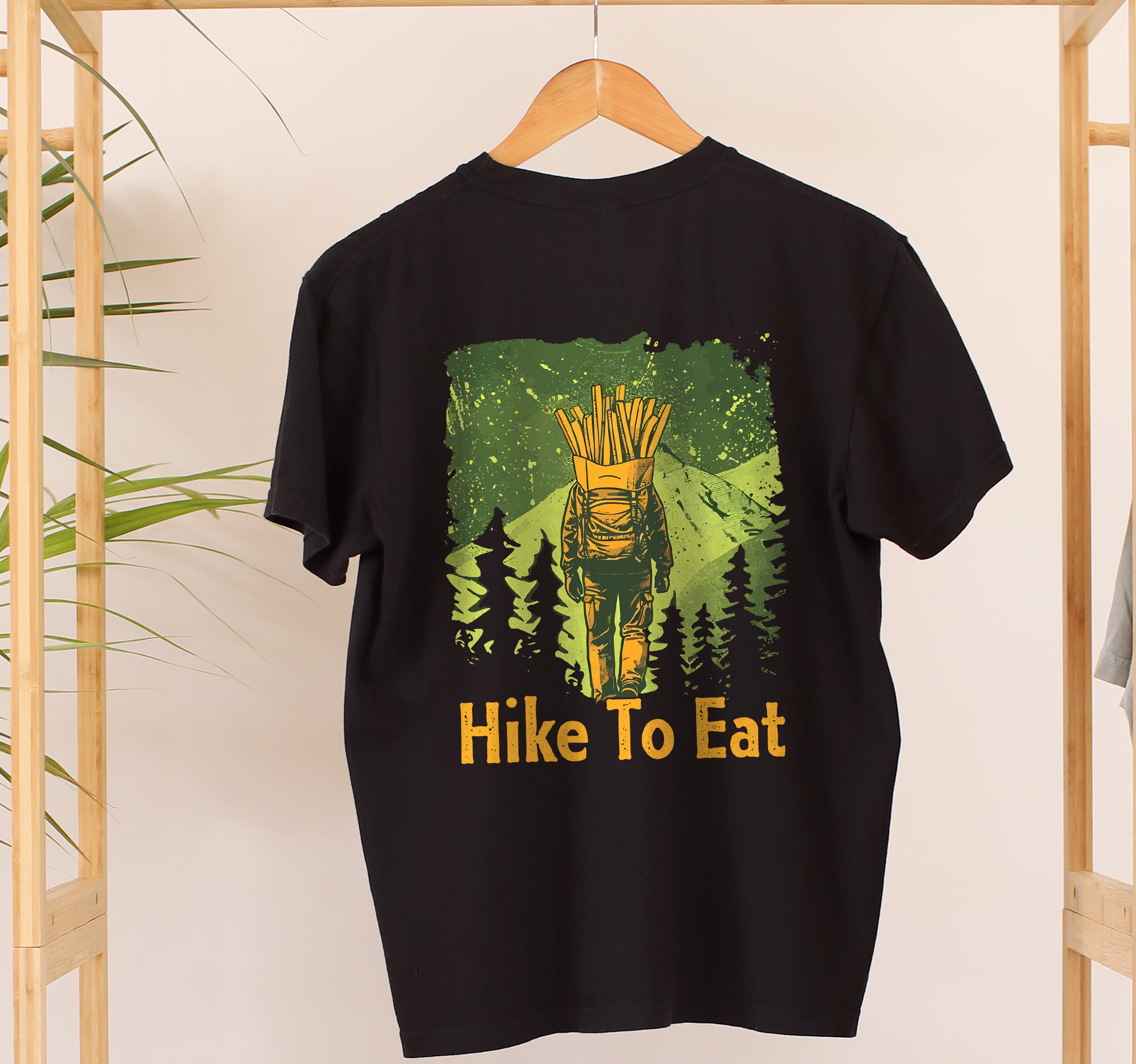 Black Hike to Eat t-shirt on a wooden hanger on back view showing large design featuring hiker with french fries backpack walking through green forest landscape with pine tree silhouettes.