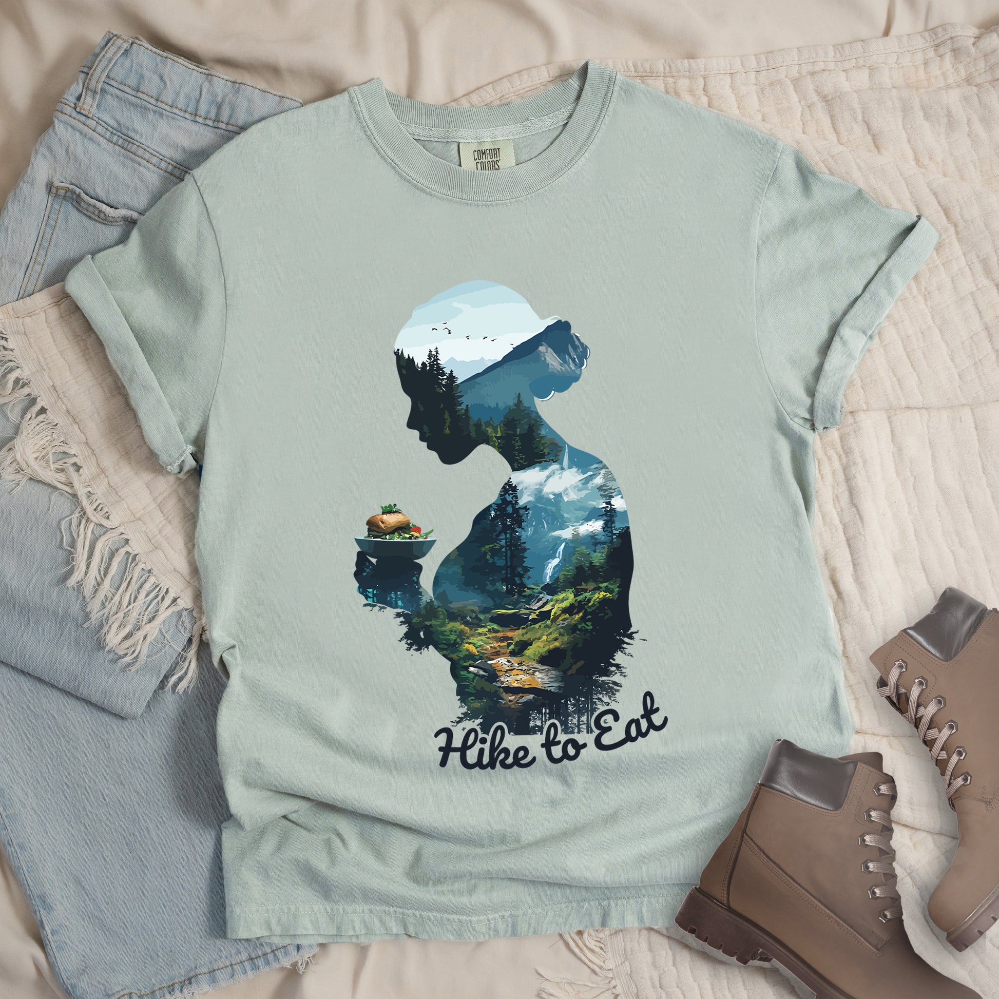 Bay (Light Gray) graphic t-shirt with 'Hike to Eat' text and 'Wilderness Within' design featuring woman's silhouette filled with mountain landscape, pine trees, and hiking trail, styled with light wash jeans and brown hiking boots