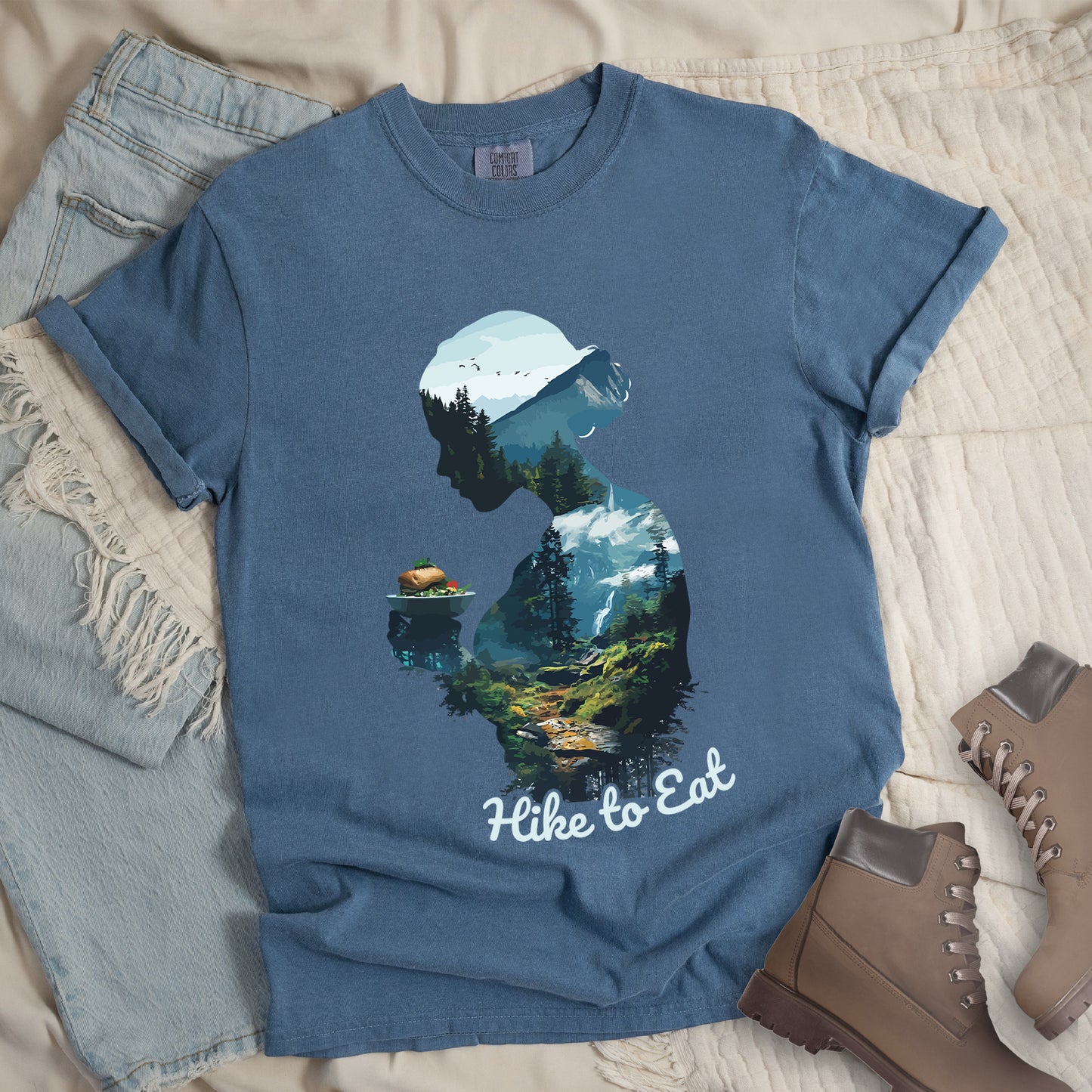 Blue Jean (Denim Blue) graphic t-shirt with 'Hike to Eat' text and 'Wilderness Within' design featuring woman's silhouette filled with mountain landscape, pine trees, and hiking trail, styled with light wash jeans and brown hiking boots