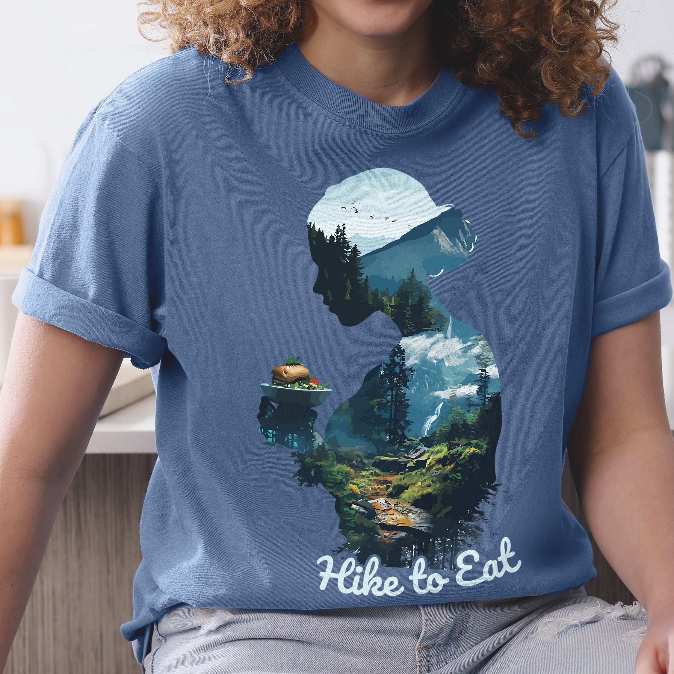 A woman wearing Wilderness Within Hike to Eat t-shirt featuring double exposure design of hiker silhouette holding plate with sandwich / burger merged with mountain forest landscape on Comfort Colors shirt in blue jean color
