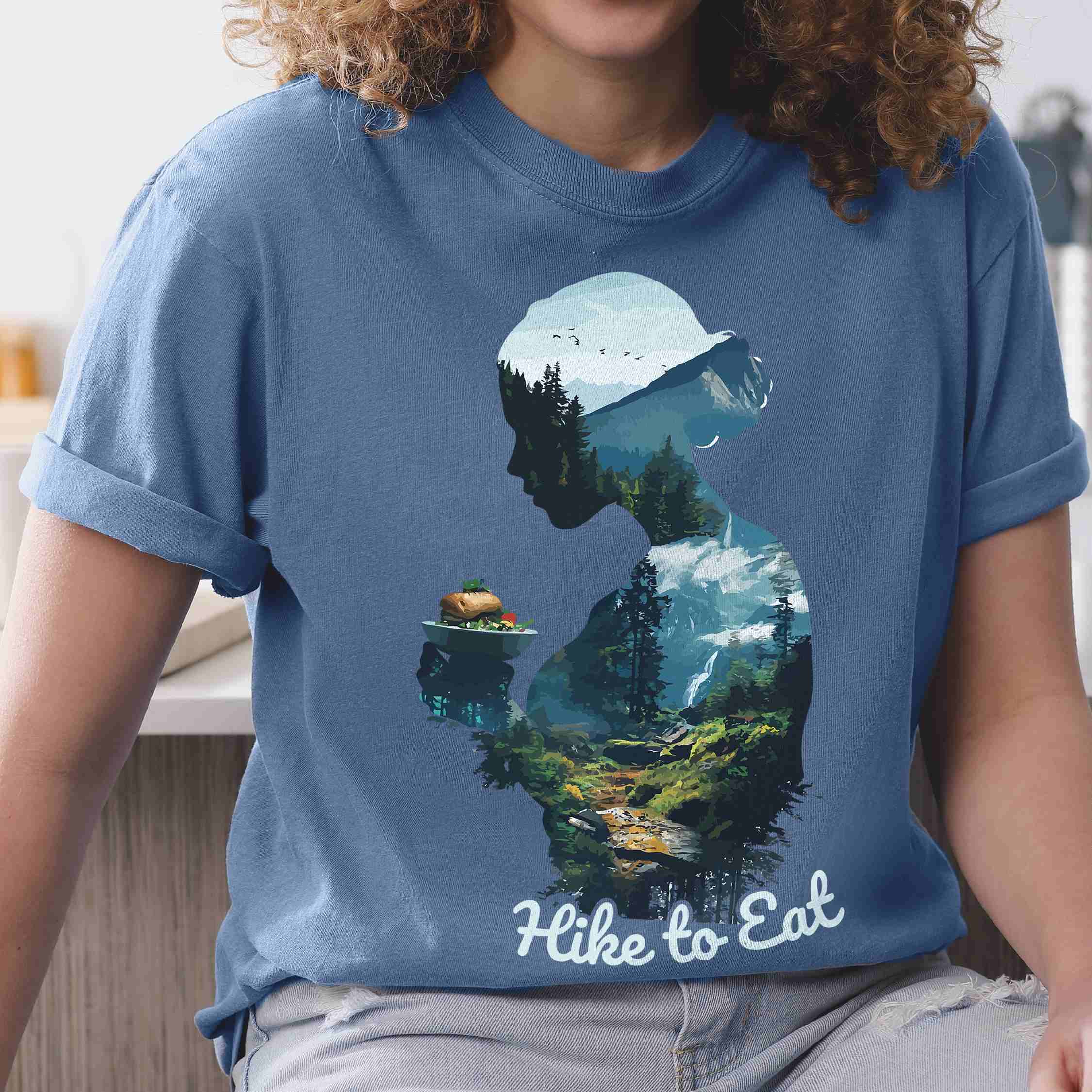 Woman wearing blue jean "Hike to Eat" colored vintage statement tee with text in cream lettering, mountains, birds, silhouette, illustrated sandwich, and backpacker for sandwich lovers.
