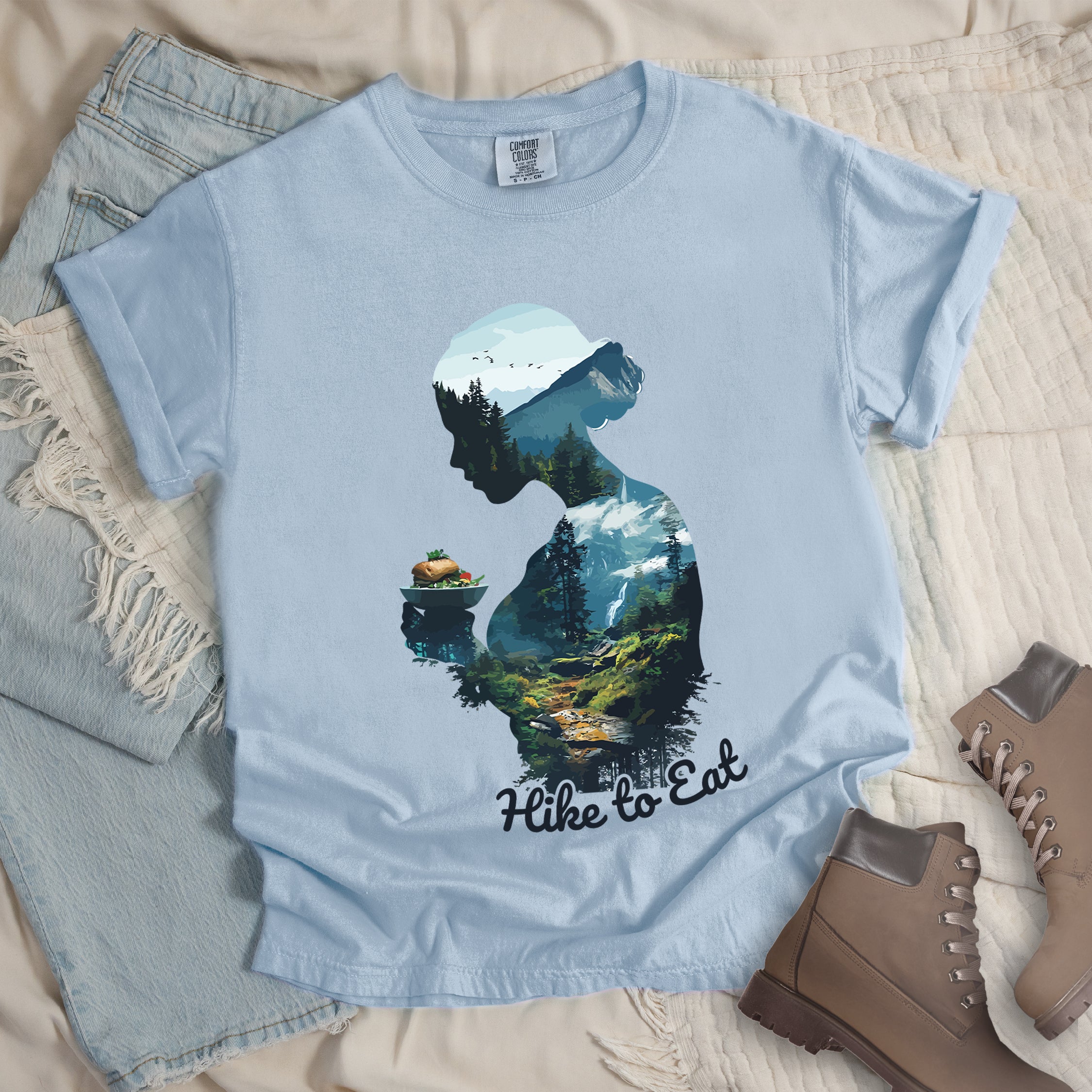 Chambray (Light Blue) graphic t-shirt with 'Hike to Eat' text and 'Wilderness Within' design featuring woman's silhouette filled with mountain landscape, pine trees, and hiking trail, styled with light wash jeans and brown hiking boots
