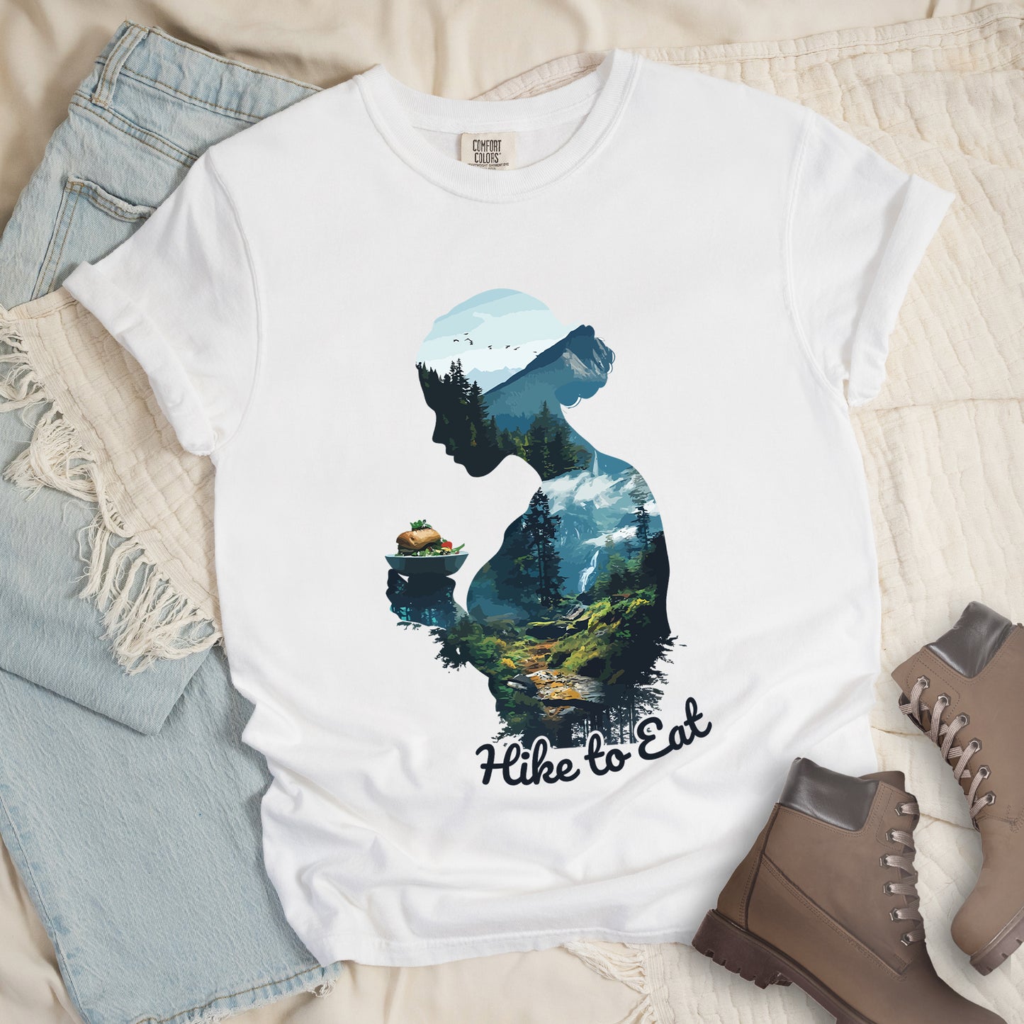 White graphic t-shirt with 'Hike to Eat' text and 'Wilderness Within' design featuring woman's silhouette filled with mountain landscape, pine trees, and hiking trail, styled with light wash jeans and brown hiking boots
