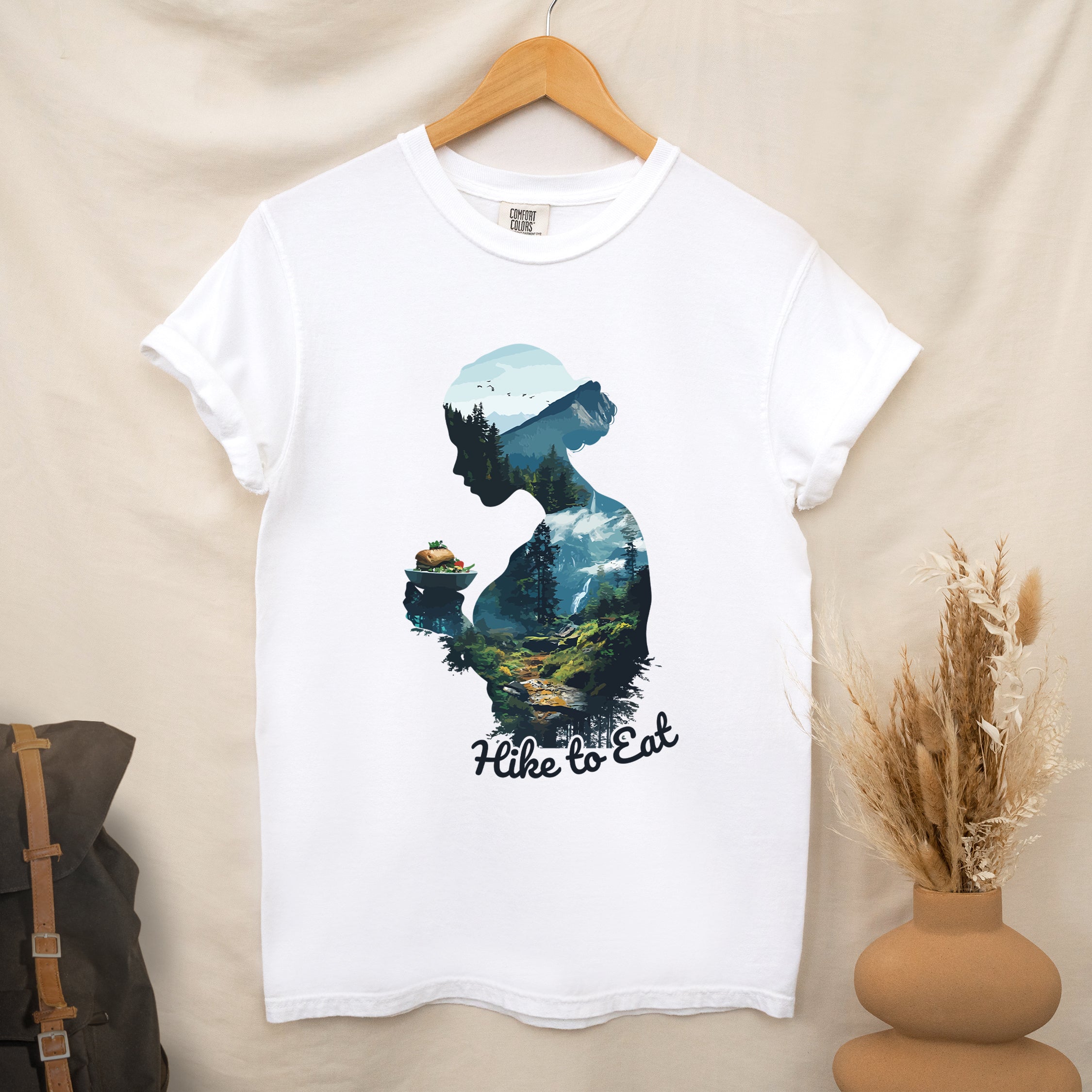 Wilderness Within Hike to Eat t-shirt featuring double exposure design of hiker silhouette holding plate with sandwich / burger merged with mountain forest landscape on Comfort Colors shirt in white hanging on a wooden hanger