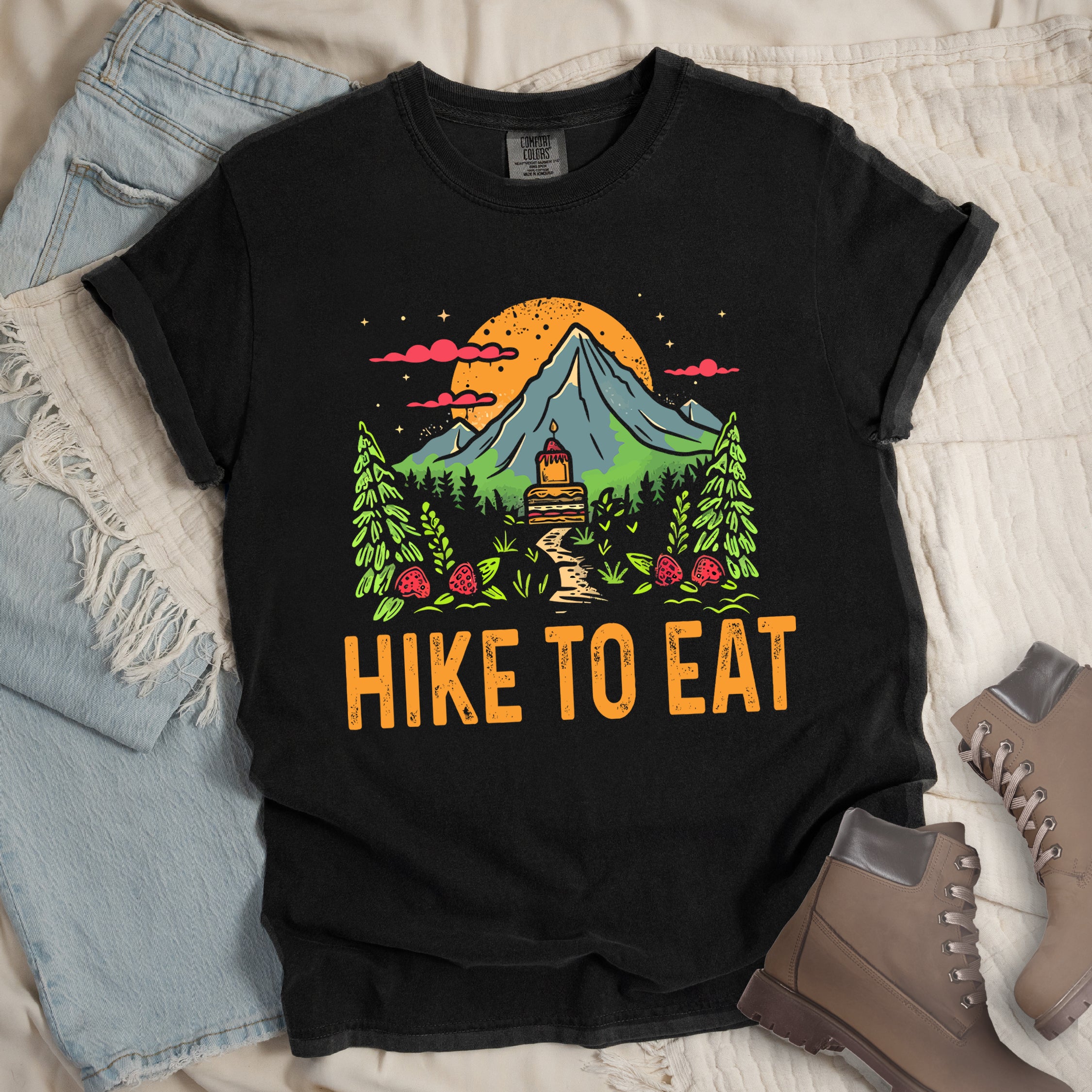 Black graphic t-shirt in flat lay view with 'Hike to Eat' text and 'Mountain Sweets' design featuring layered cake in mountain landscape with orange sunset, pine trees, mushrooms, and starry sky.