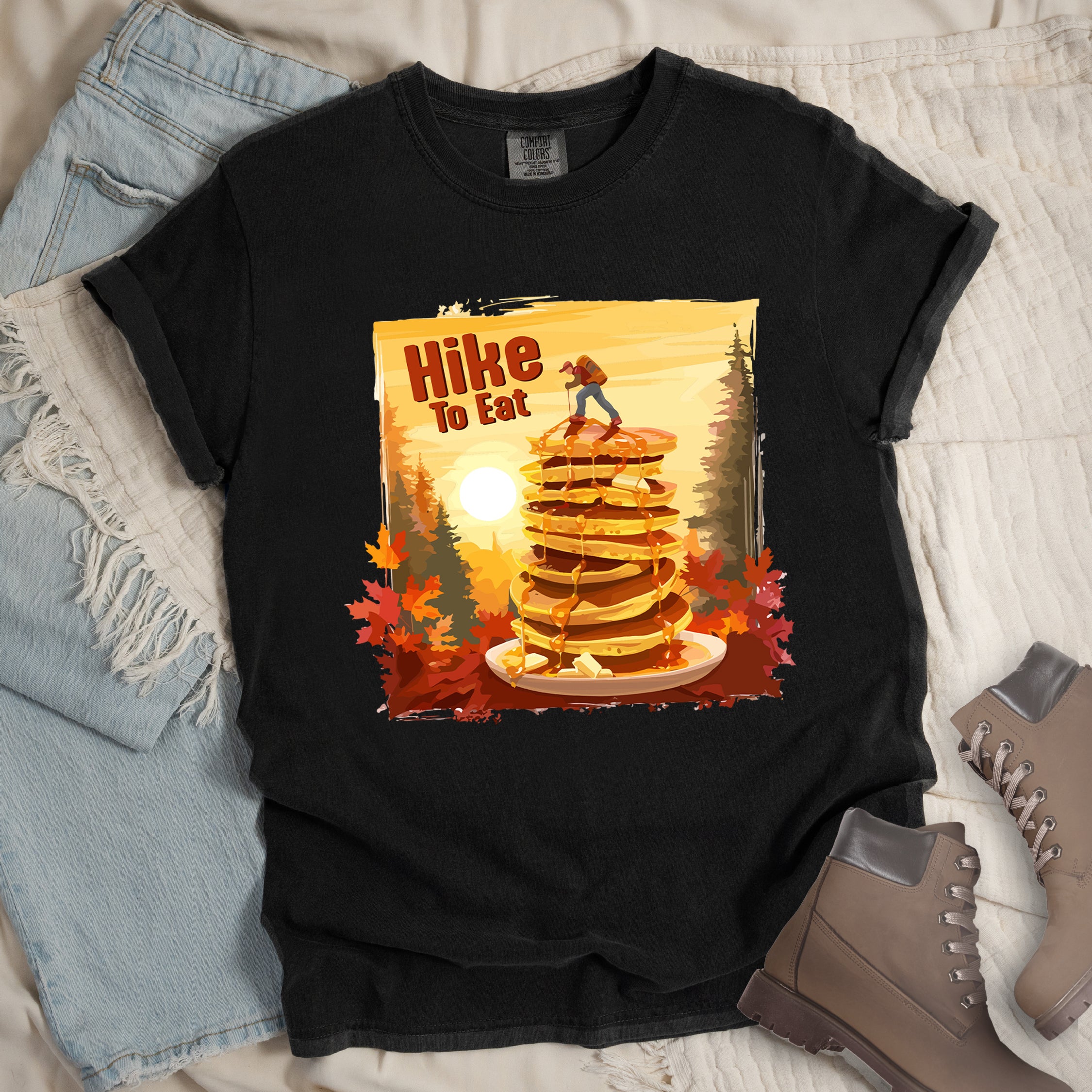 Black graphic t-shirt with 'Hike to Eat' text and 'Pancake Summit' design featuring tall stack of pancakes with hiker figure on top, set against autumn forest landscape with orange sunset, styled with light wash jeans and brown hiking boots.
