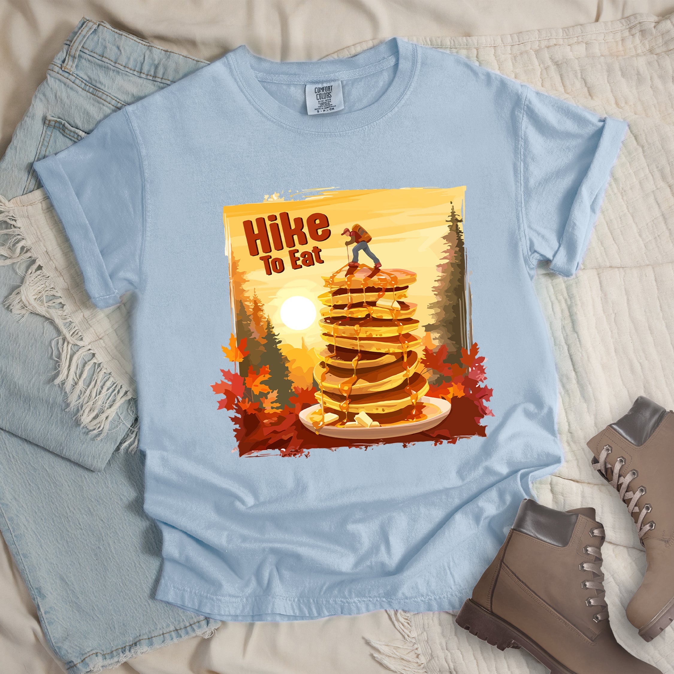 Chambray (Light Blue) graphic t-shirt with 'Hike to Eat' text and 'Pancake Summit' design featuring tall stack of pancakes with hiker figure on top, set against autumn forest landscape with orange sunset, styled with light wash jeans and brown hiking boots.