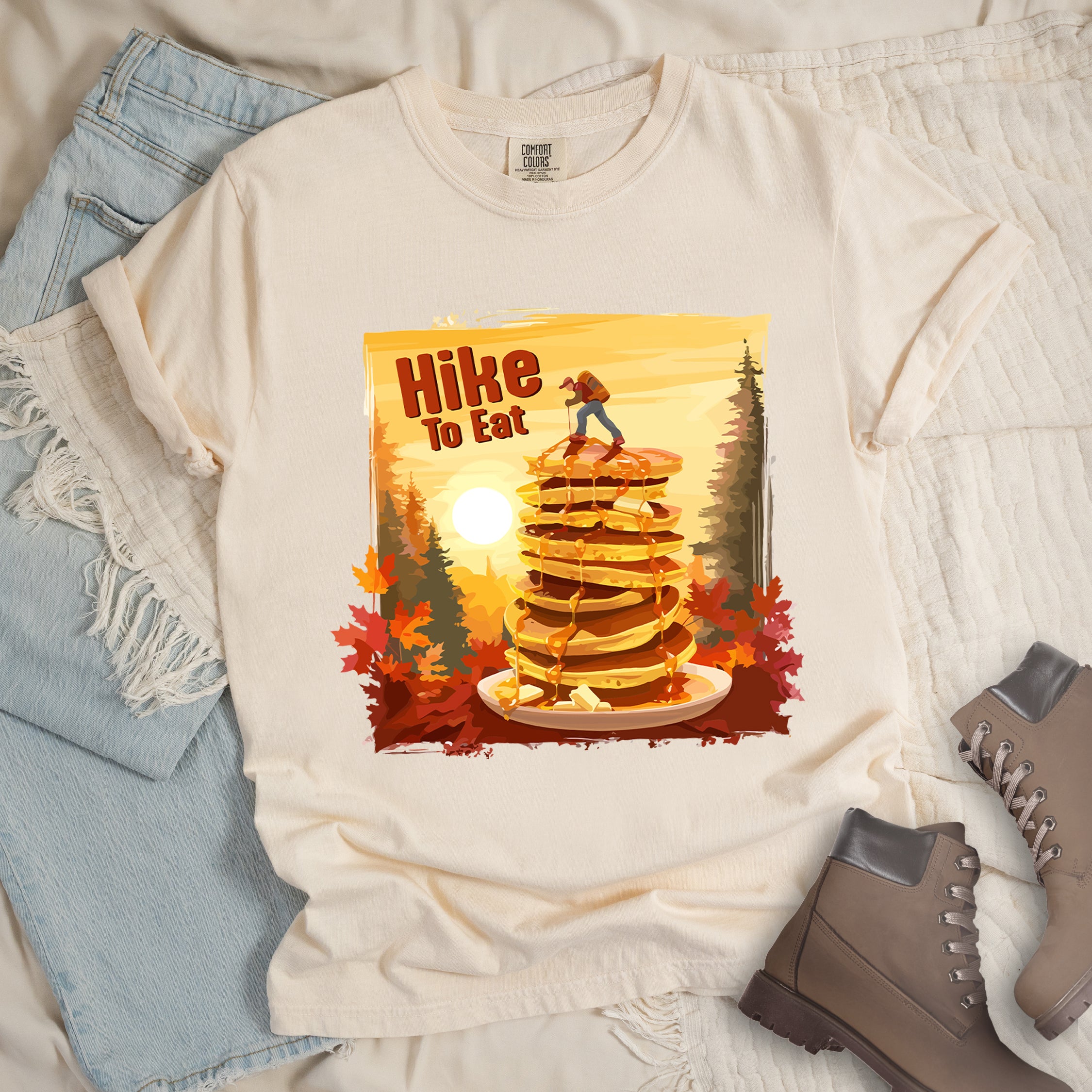 Ivory (Cream White) graphic t-shirt with 'Hike to Eat' text and 'Pancake Summit' design featuring tall stack of pancakes with hiker figure on top, set against autumn forest landscape with orange sunset, styled with light wash jeans and brown hiking boots