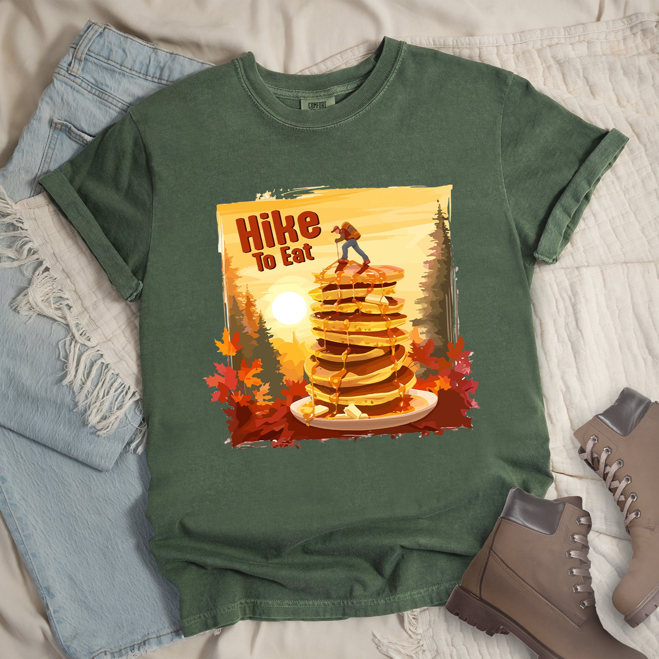 Moss (Dark Green) graphic t-shirt with 'Hike to Eat' text and 'Pancake Summit' design featuring tall stack of pancakes with hiker figure on top, set against autumn forest landscape with orange sunset, styled with light wash jeans and brown hiking boots