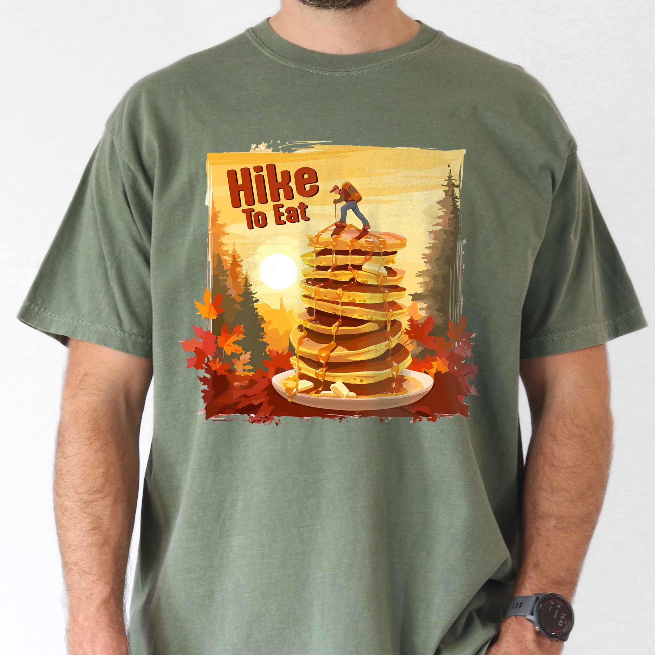 Man wearing moss "Hike to Eat" colored vintage statement tee with text in bold lettering, mountains, birds, silhouette, illustrated stack of pancake, and perfect for breakfast lovers.