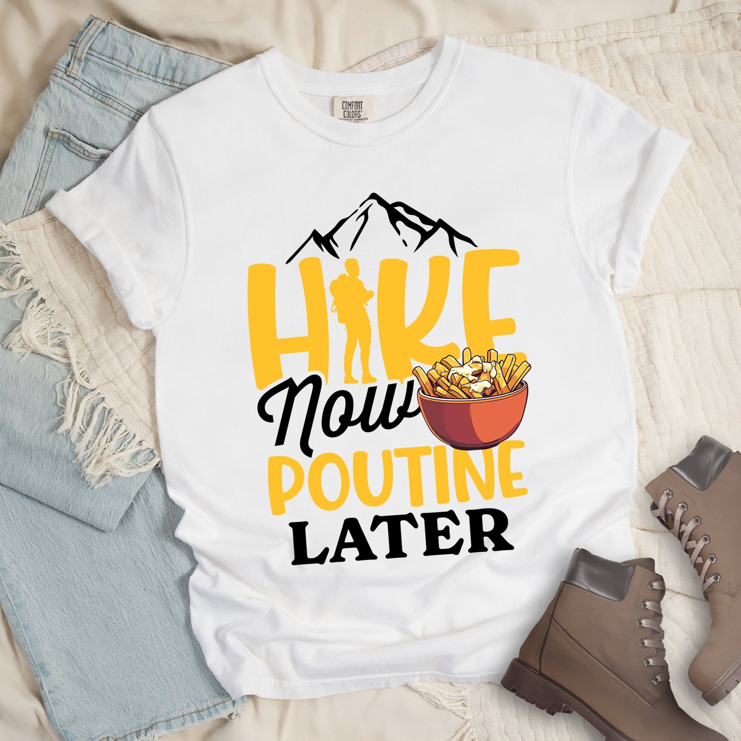 White t-shirt with "HIKE NOW POUTINE LATER" text design. Black mountain silhouettes appear above yellow "HIKE" letters, with a hiker silhouette replacing the "I". "Now" appears in black script, "POUTINE" in yellow capital letters, and "LATER" in black. An illustration of a bowl of poutine with french fries, cheese curds and gravy appears next to "Now" and  above "POUTINE". Features a classic crew neck and casual rolled sleeves, styled alongside light wash denim and brown hiking boots. 