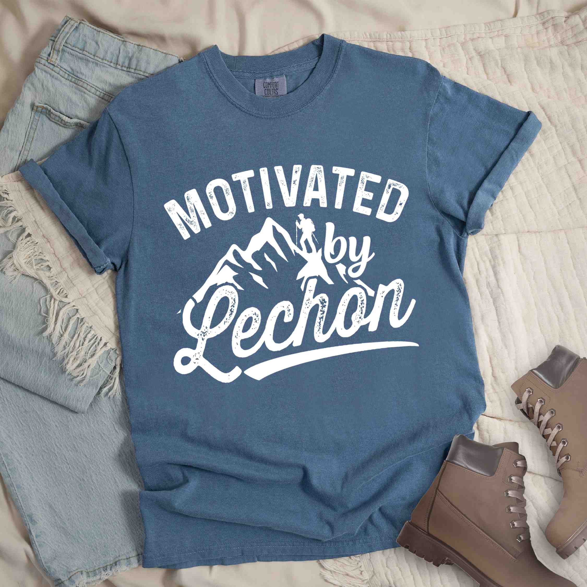 Blue jean colored graphic t-shirt with 'Motivated by Lechon' text in white lettering featuring mountain silhouettes and hiker illustration