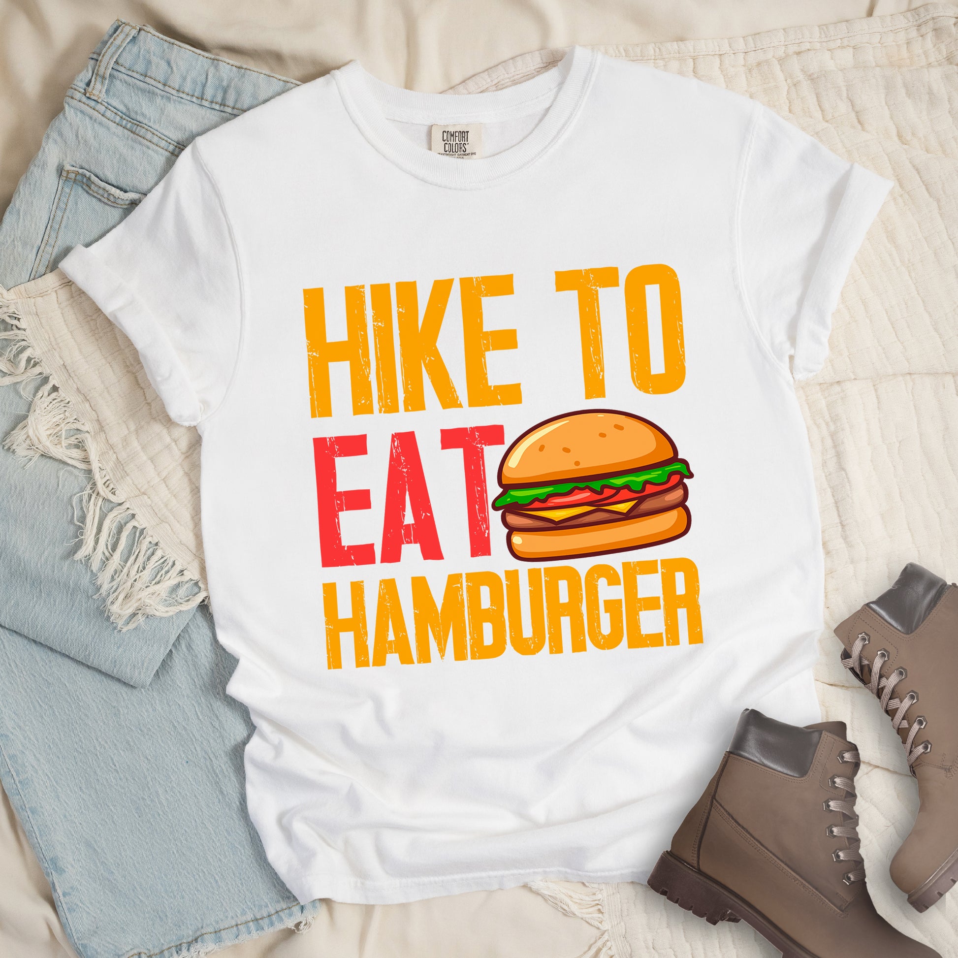 White t-shirt with distressed text reading "HIKE TO EAT HAMBURGER" arranged in three lines. "HIKE TO" appears in yellow at the top, "EAT" in red in the middle with a colorful hamburger illustration next to it, and "HAMBURGER" in yellow at the bottom. The hamburger graphic shows a bun, meat patty, cheese, lettuce, and tomato. Features a classic crew neck and casual rolled sleeves, styled alongside light wash denim and brown hiking boots. 