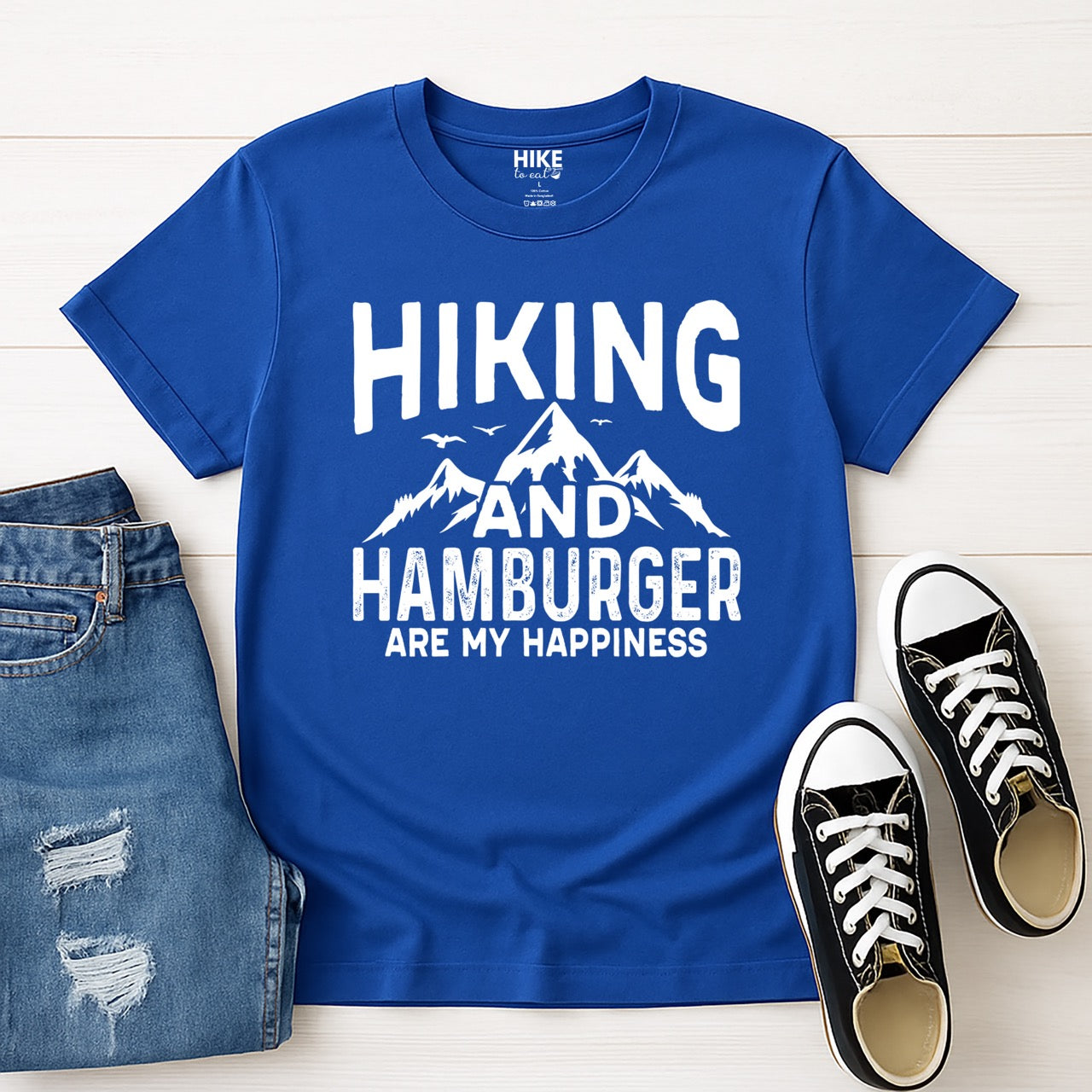  royal (blue) Hiking and Hamburger are my Happiness statement tee with distressed bold typography, mountain peaks, flying birds, and pine trees for burger-loving outdoor enthusiasts and hikers - layed flat with jeans and shoes
