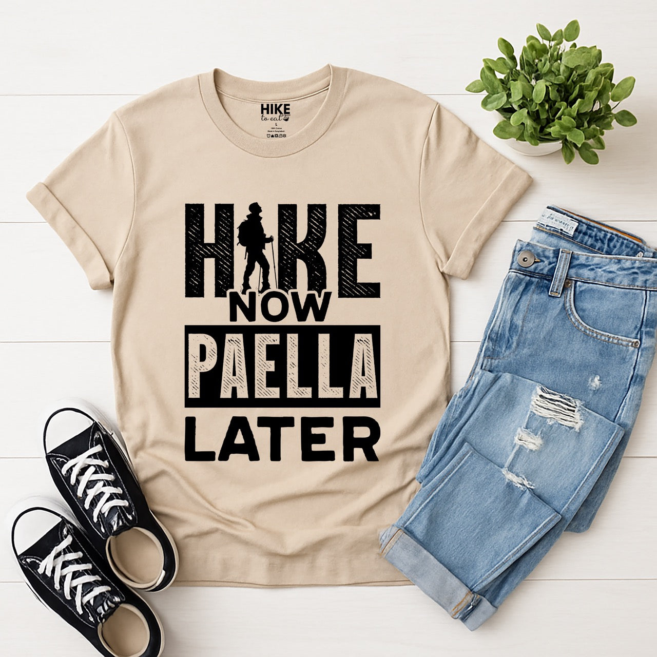 sand (beige) Hike Now Paella Later statement tee with bold distressed typography and hiker silhouette for Spanish food-loving outdoor enthusiasts and hikers with jeans and shoes