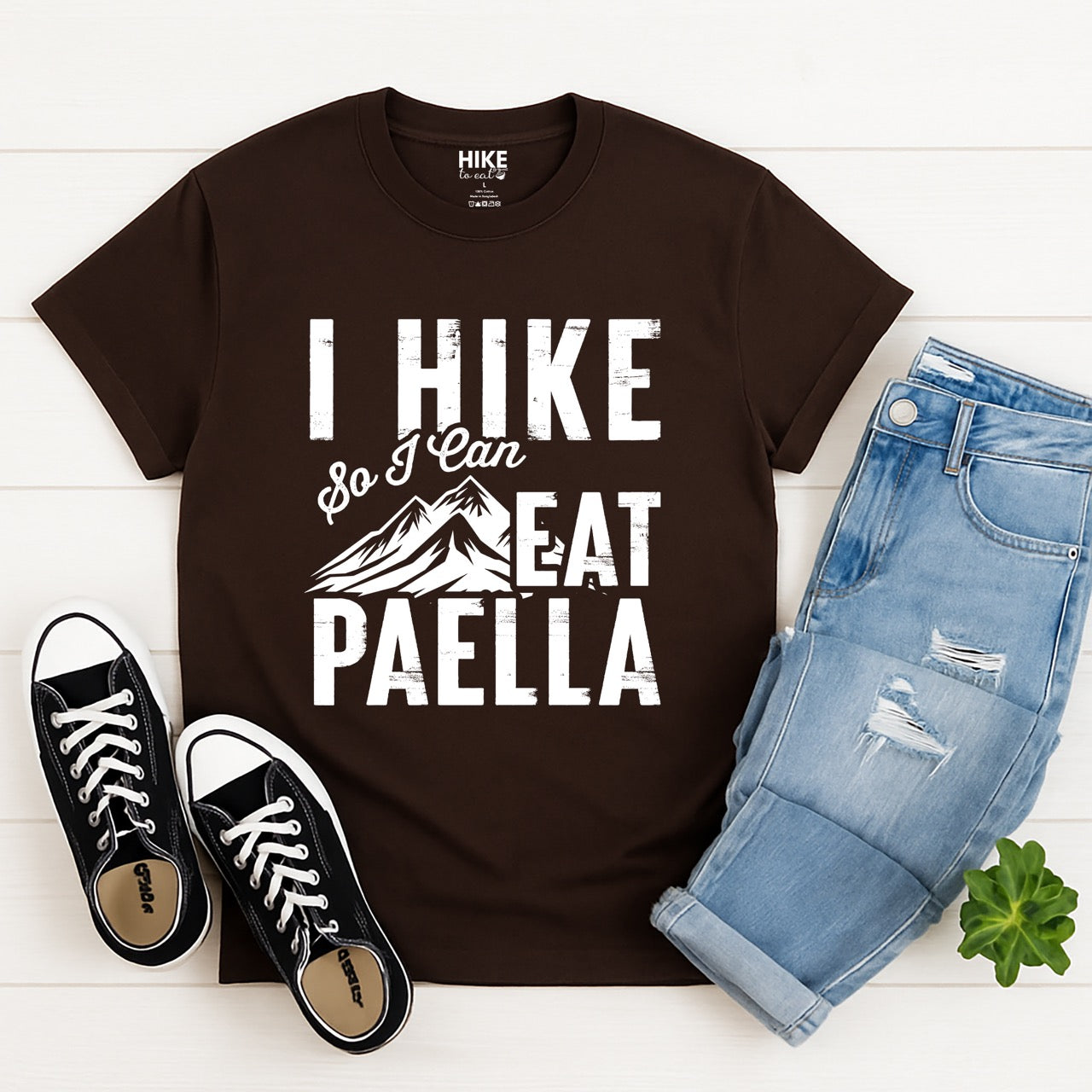 chocolate brown Hike Now Paella Later statement tee with bold distressed typography and hiker silhouette for Spanish food-loving outdoor enthusiasts and hikers with jeans and shoes