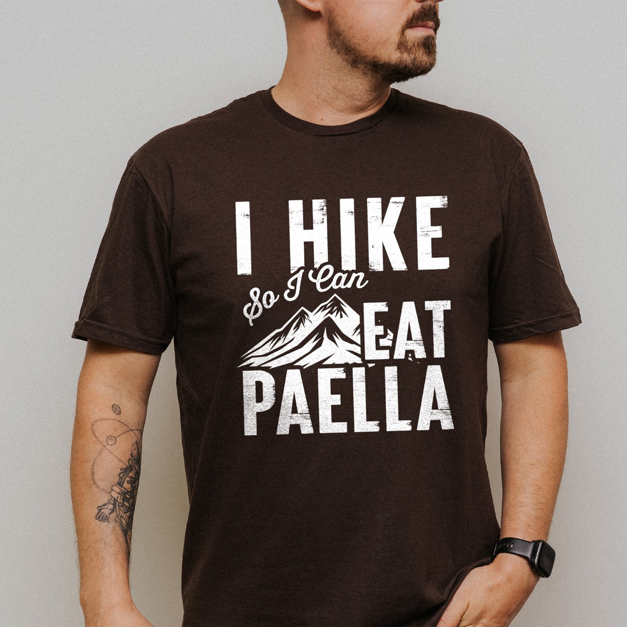 man wearing chocolate brown Hike Now Paella Later statement tee with bold distressed typography and hiker silhouette for Spanish food-loving outdoor enthusiasts and hikers
