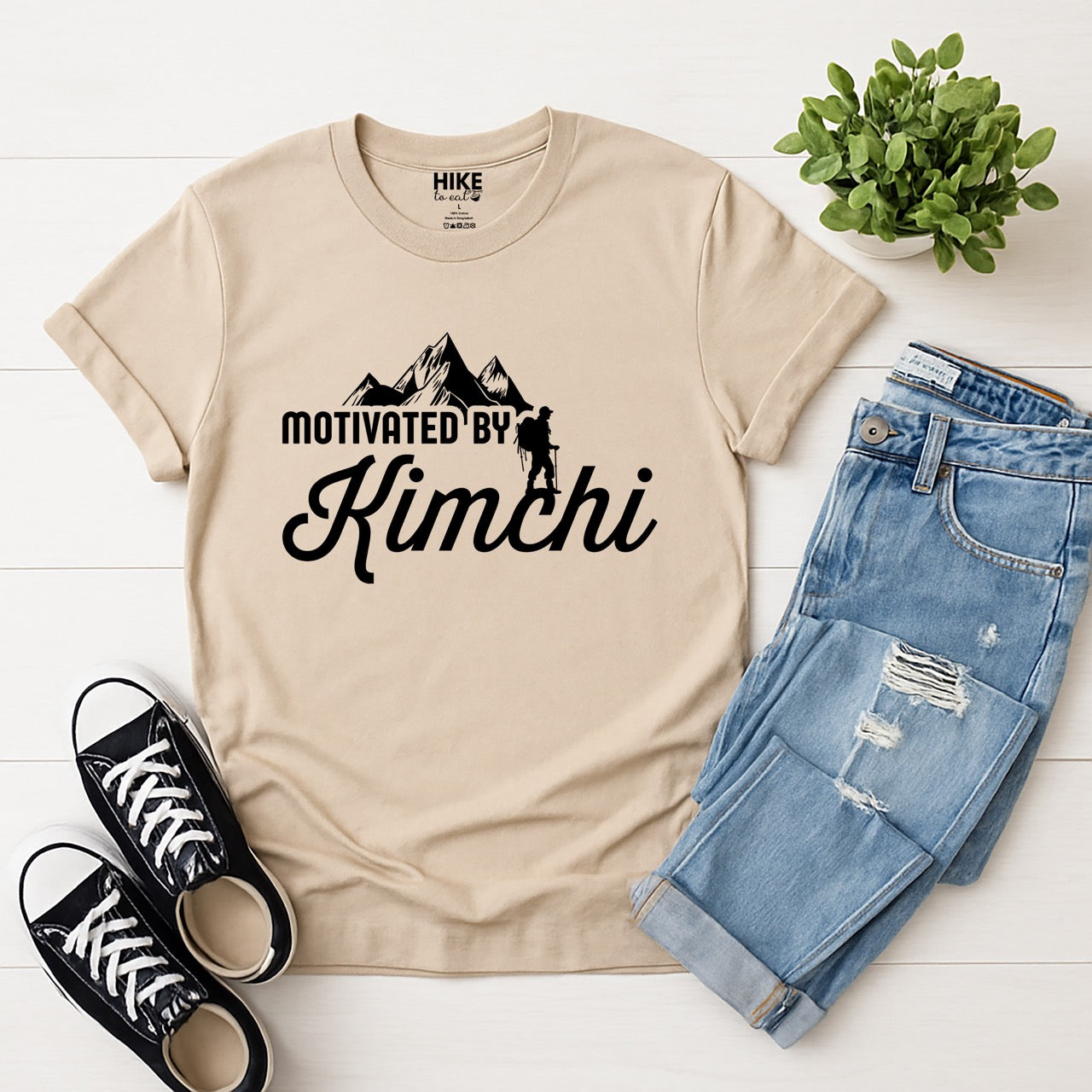 sand (beige) colored Motivated by Kimchi statement tee with bold typography, elegant script lettering, mountain peaks, and hiker silhouette for Korean food-loving outdoor enthusiasts, foodies and hikers laid flat with jeans and shoes