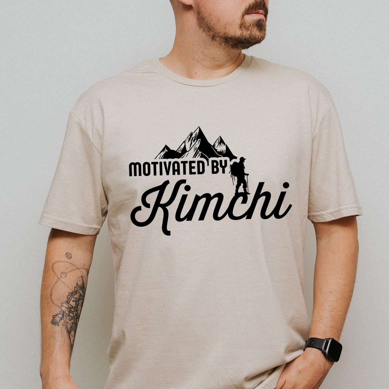 man wearing sand (beige) colored Motivated by Kimchi statement tee with bold typography, elegant script lettering, mountain peaks, and hiker silhouette for Korean food-loving outdoor enthusiasts, foodies and hikers