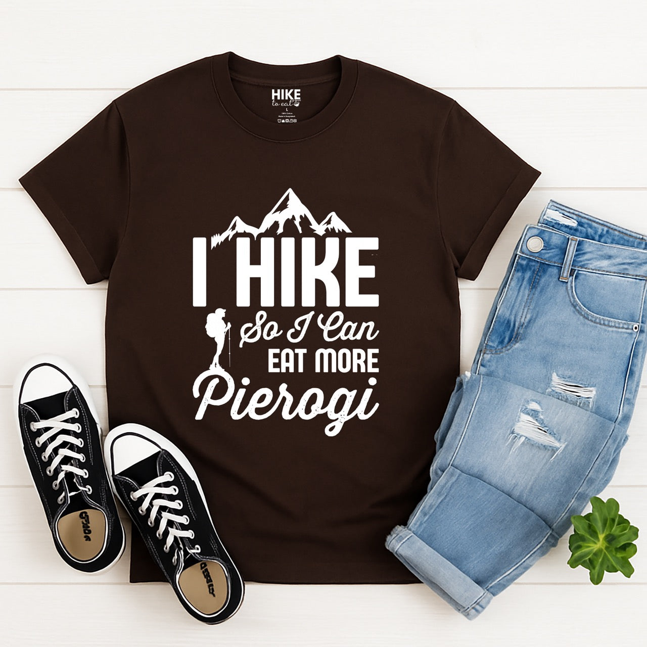 Chocolate brown colored I Hike so I can eat more Pierogi statement tee with distressed bold typography, mountain peaks, pine trees, and hiker silhouette for Polish food-loving outdoor enthusiasts, hikers and foodies - laid flat with jeans and shoes