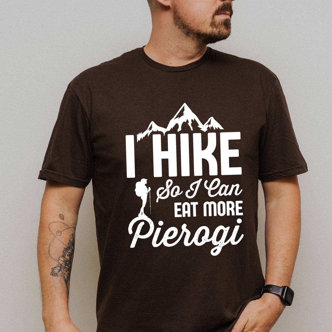 Man wearing Chocolate brown colored I Hike so I can eat more Pierogi statement tee with distressed bold typography, mountain peaks, pine trees, and hiker silhouette for Polish food-loving outdoor enthusiasts, hikers and foodies