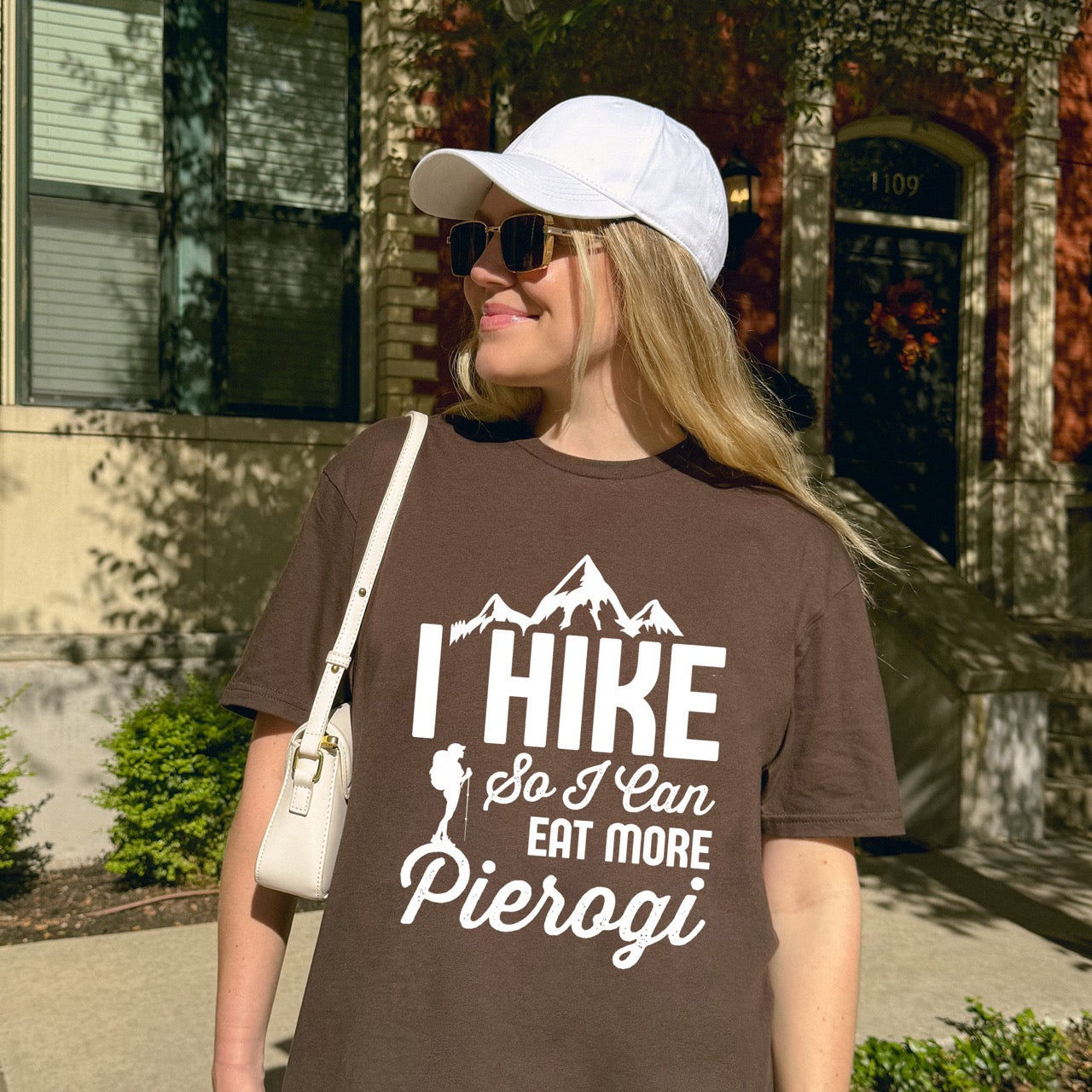 Woman wearing Chocolate brown colored I Hike so I can eat more Pierogi statement tee with distressed bold typography, mountain peaks, pine trees, and hiker silhouette for Polish food-loving outdoor enthusiasts, hikers and foodies