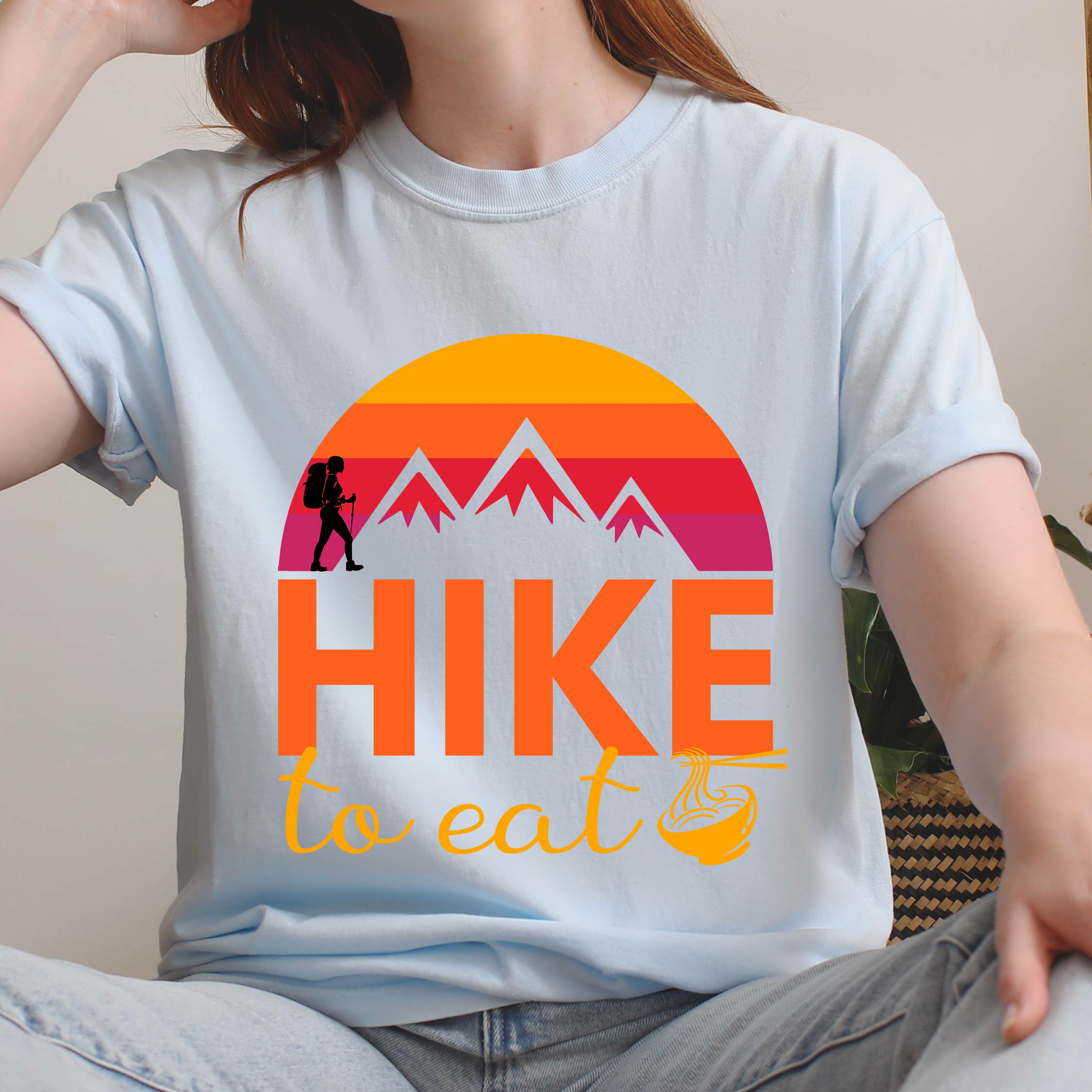 Woman wearing chambray "Hike to Eat" colored vintage tee with bold typography, mountains, sunrise, and backpacker to savor each step along the trail.
